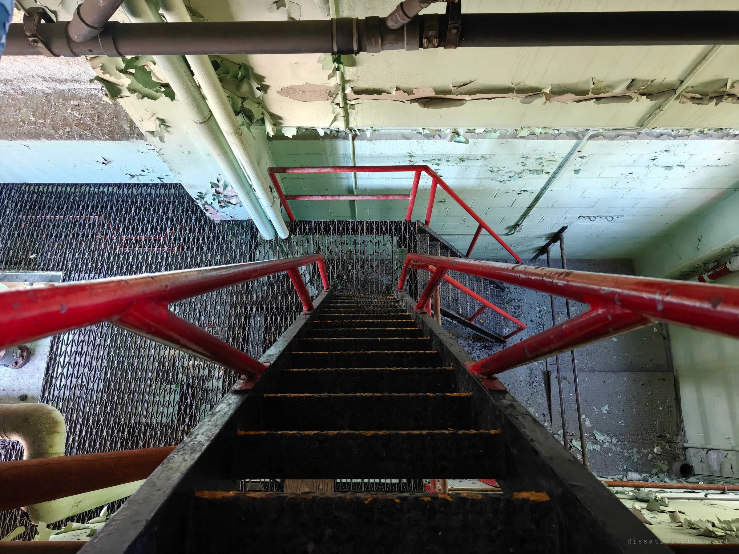 Looking straight down a steep black stair with red rails into a lower landing of mesh grating and flaking green walls. Paint chips cover the floor below.