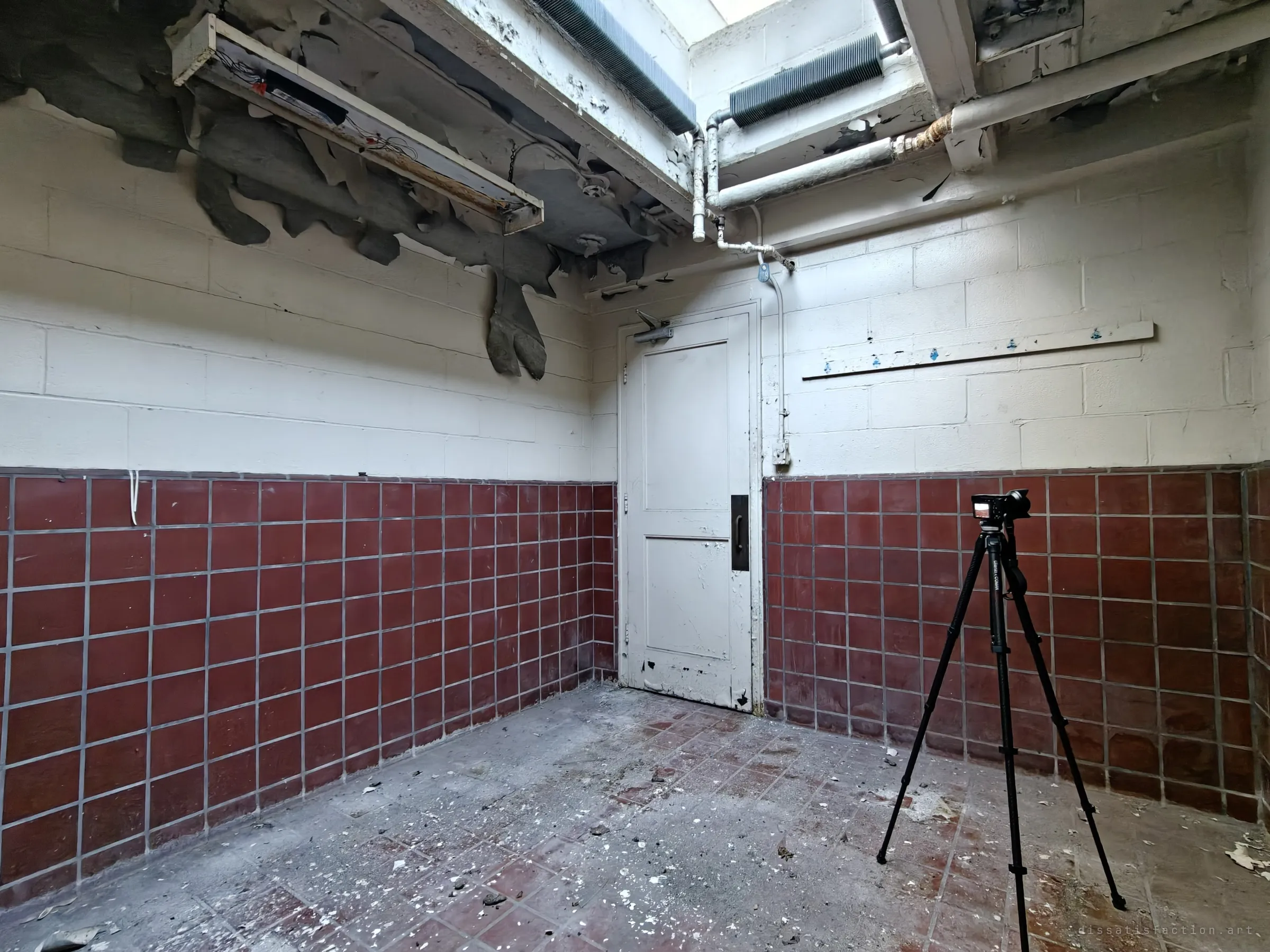 A small room with oxblood tile wainscot, a closed white door and a broken light fixture sagging from the ceiling, with a tripod-mounted camera in the corner. Light falls in from a shaft overhead.
