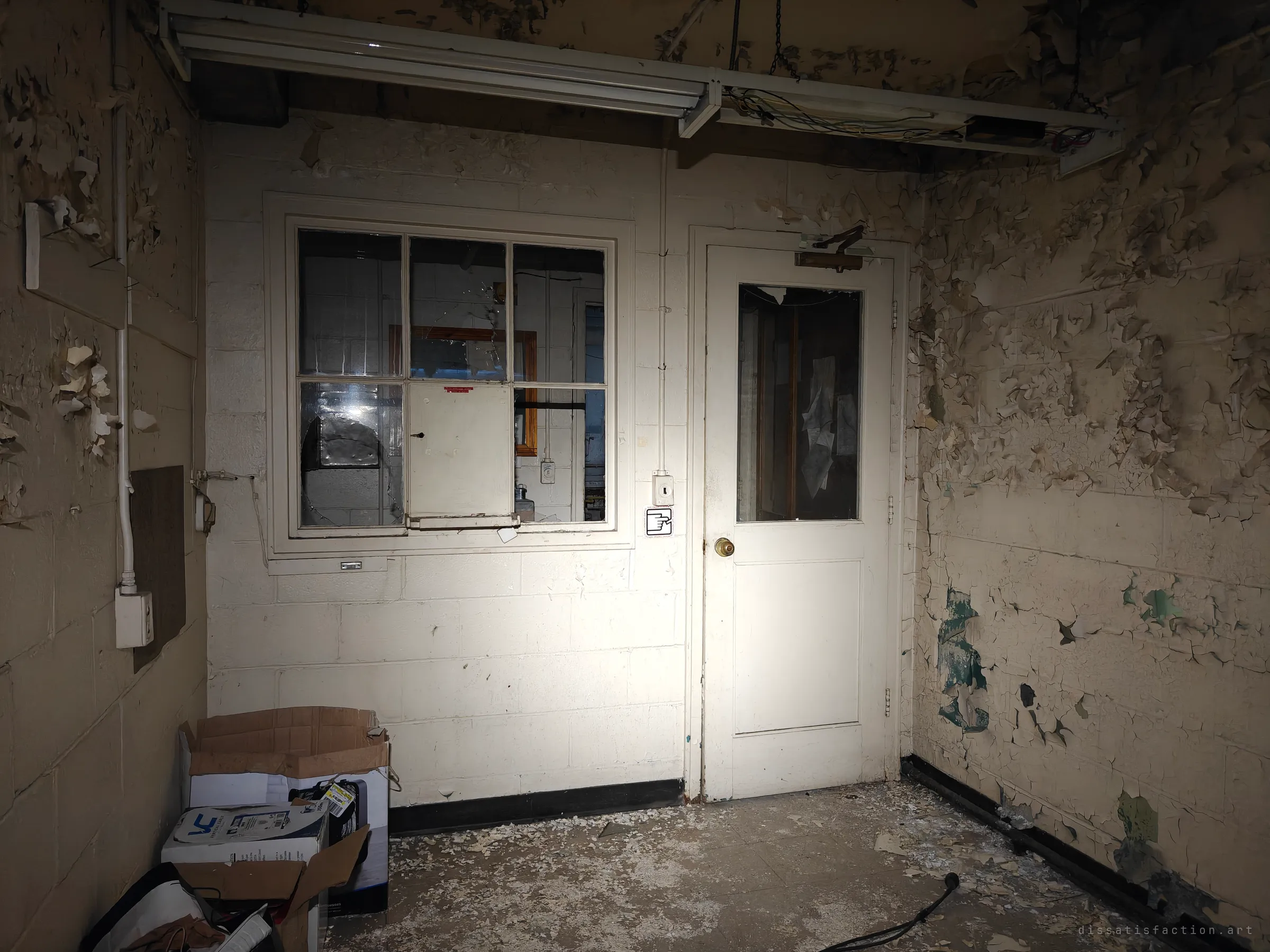 A flashlit anteroom with a service window and a half-glazed door into an office where papers still hang inside. Boxes sit abandoned in the corner under a gutted light fixture.