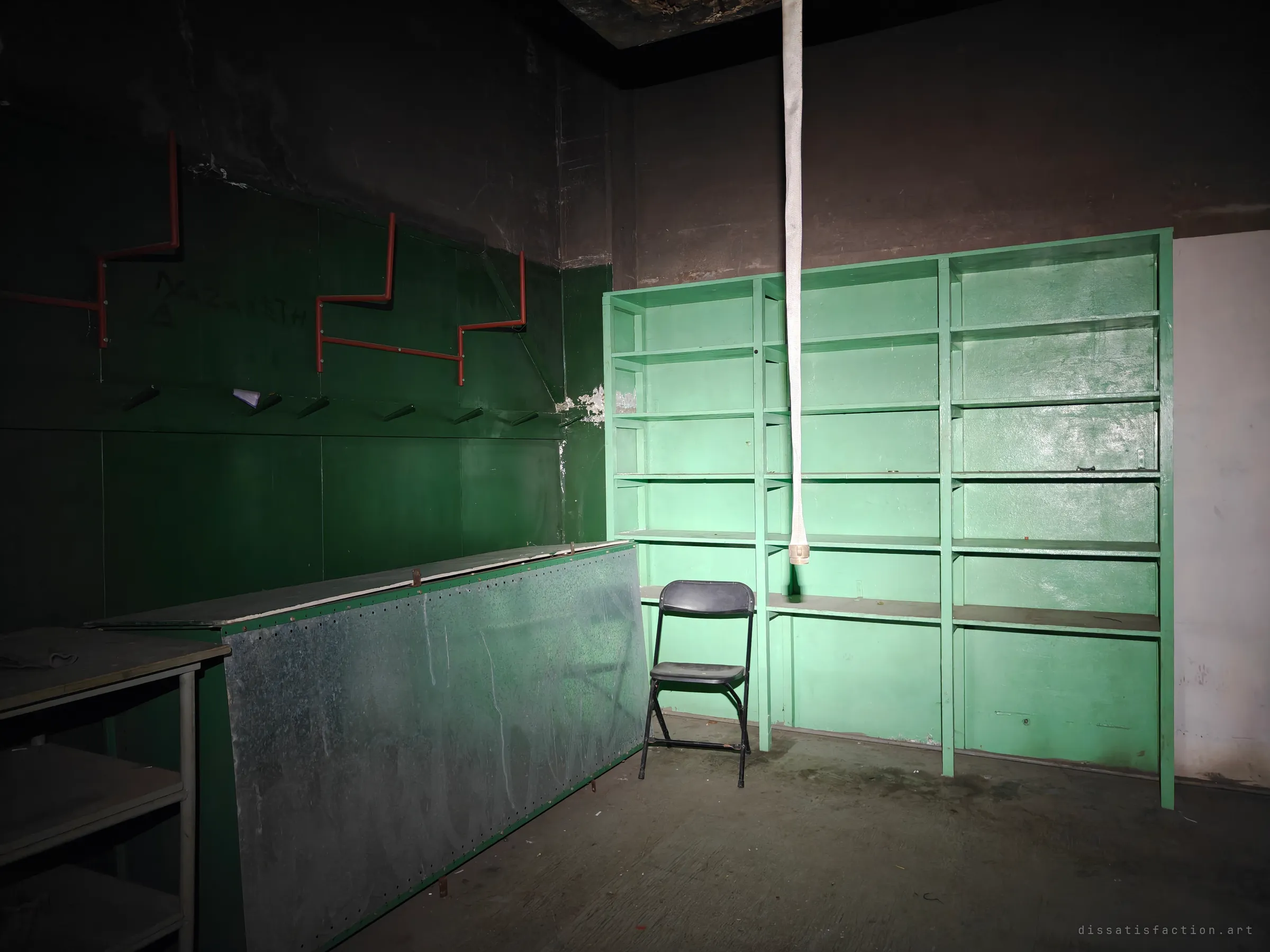 A dark locker room with bright green empty shelving, a lone folding chair and a fire hose hanging from the ceiling. Red zigzag coat racks run along the shadowed wall.