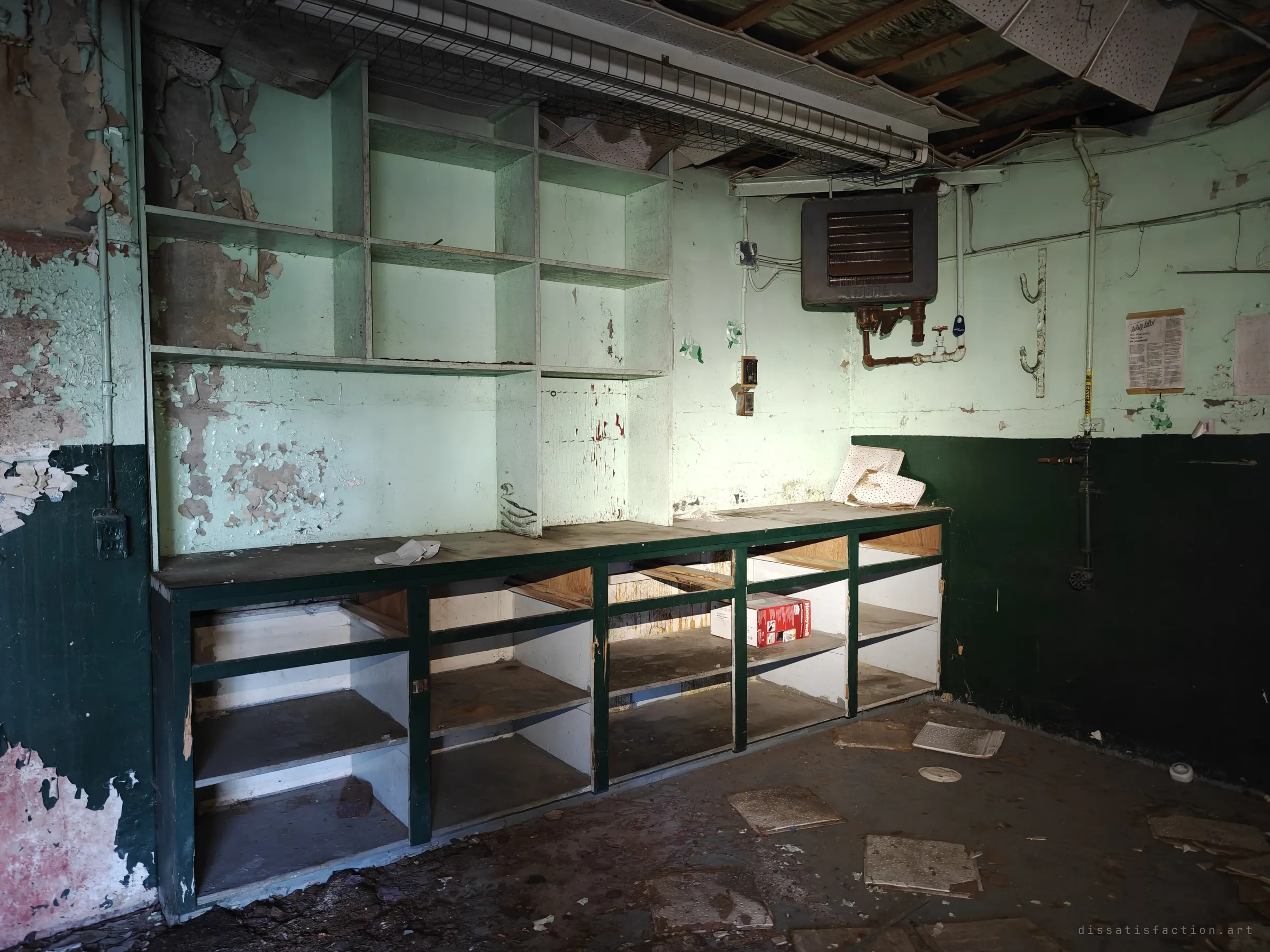 An emptied workshop with pigeonhole shelving over a dark green counter, a rusting unit heater and an old safety poster on the wall. Ceiling tiles have dropped to the floor.