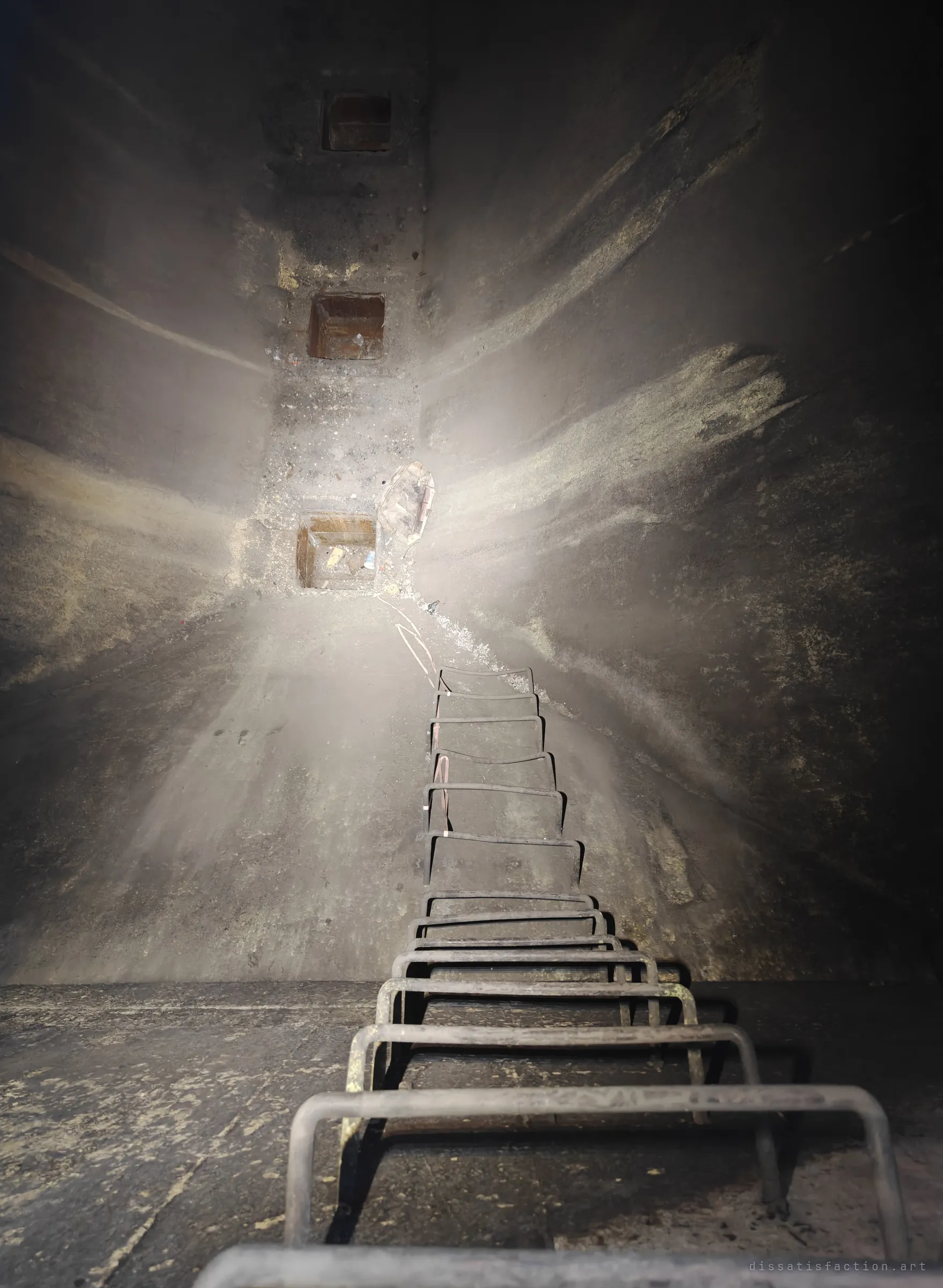 Looking up a soot-blackened shaft along a steel rung ladder toward square openings lit by a torch beam. The narrow climb shows how deep into the plant's flues the space goes.