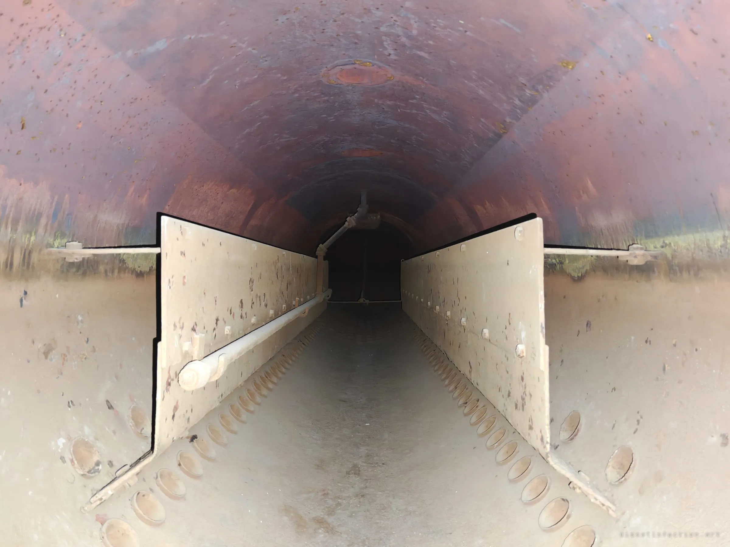 The inside of a large boiler drum, a rusted curved shell with rows of tube holes and two baffle plates running to a dark end. The symmetry of the drum draws the eye straight down its length.