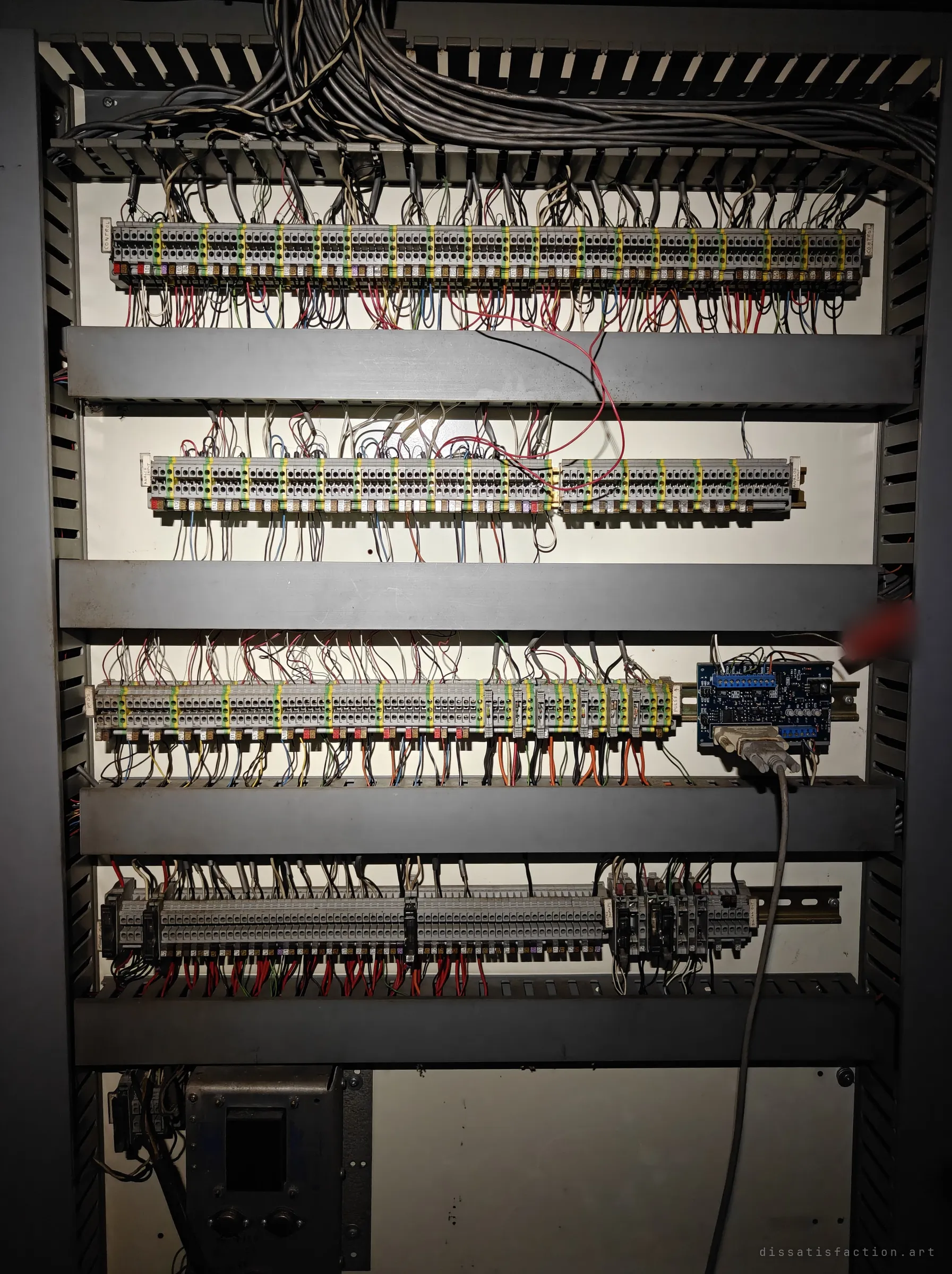 An open control cabinet packed with rows of terminal blocks, bundled wiring and a small blue circuit board with a serial cable. The dense wiring shows how much of the plant was still automated.
