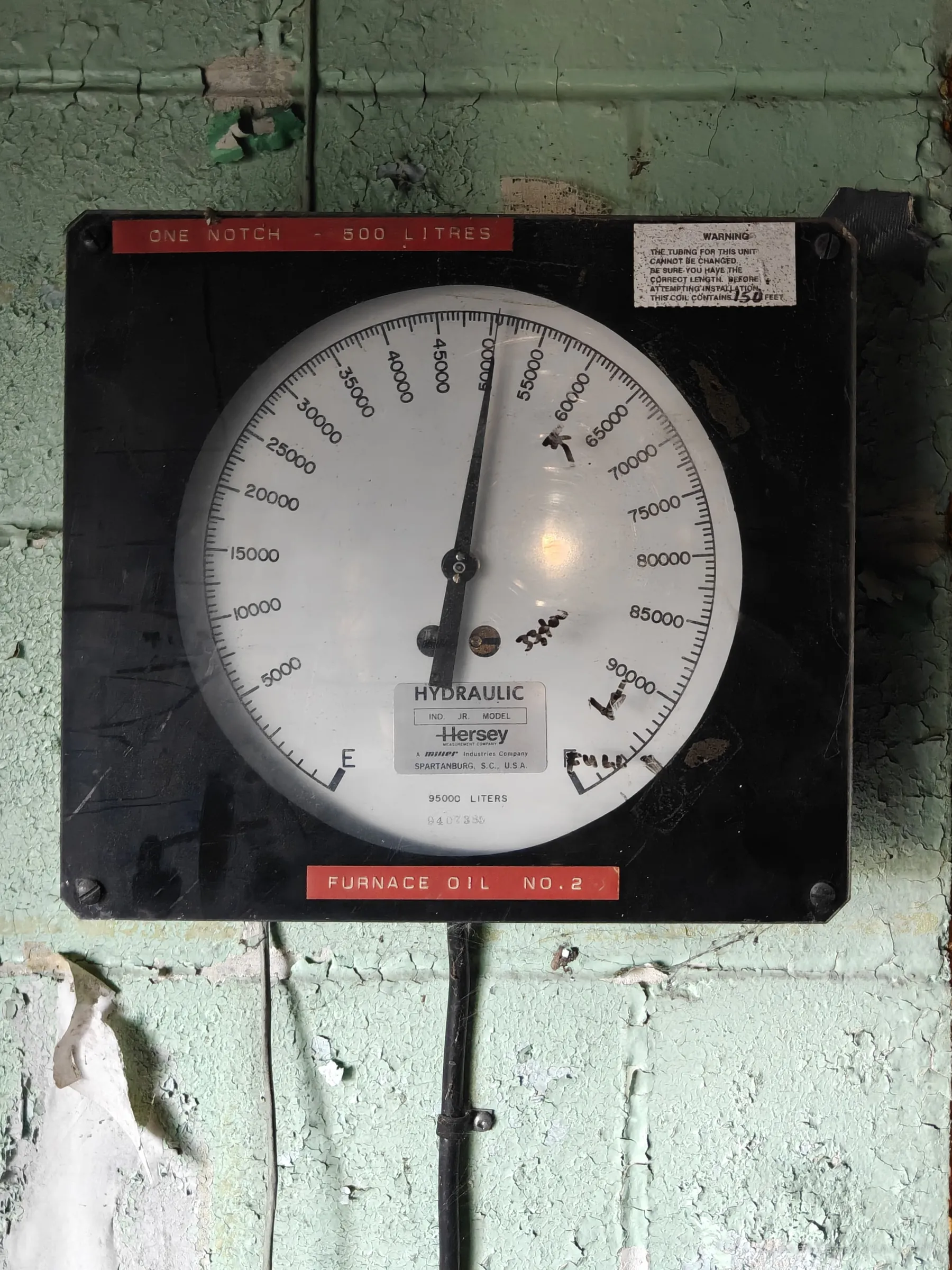 A large hydraulic tank gauge for furnace oil on a flaking green block wall, its needle resting near the half-full mark with handwritten ticks on the dial. It hints at fuel still sitting in the tanks when the plant went quiet.
