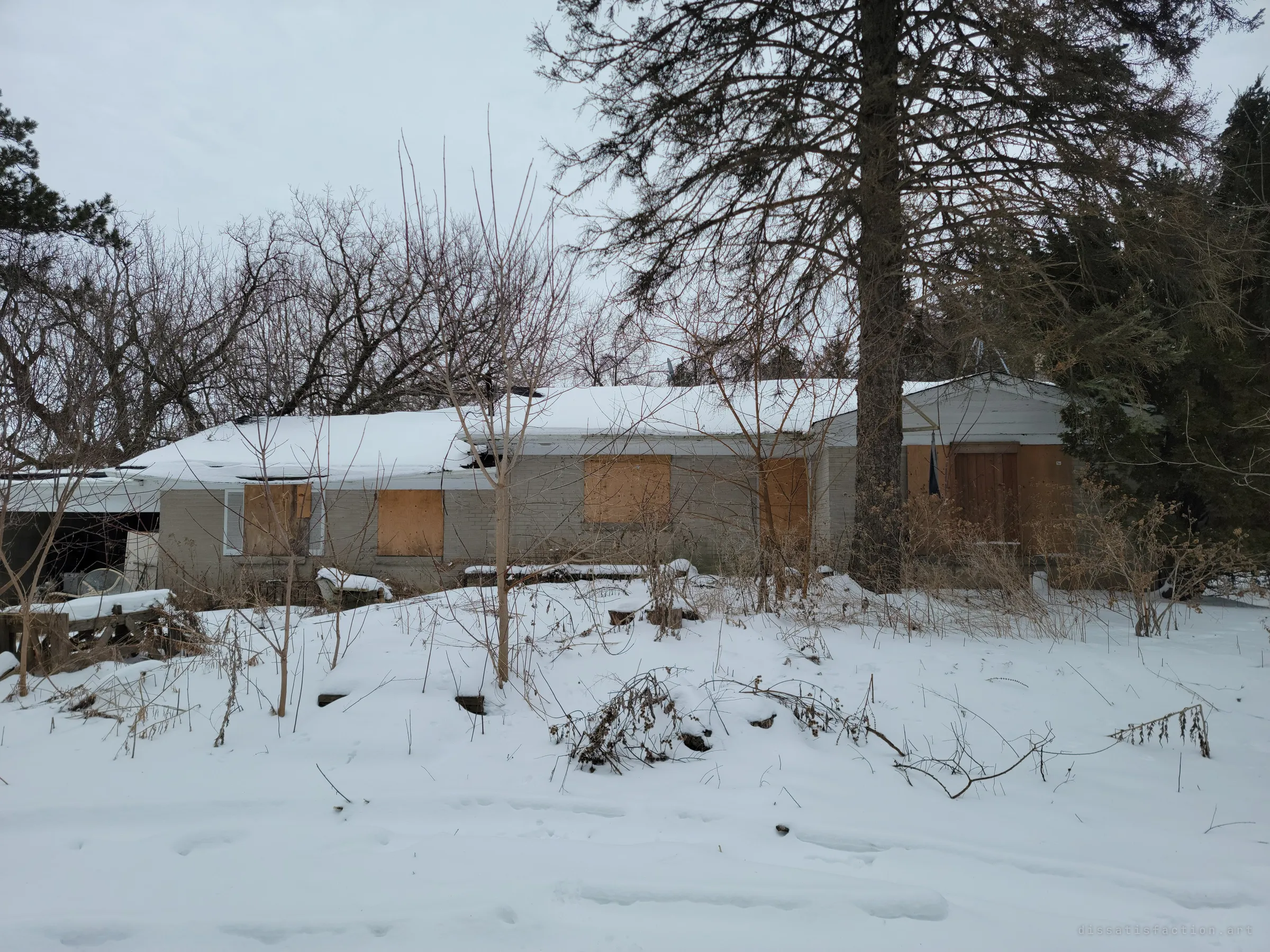 A single-storey brick bungalow sits under snow with every window boarded over in plywood. Bare trees and a tall pine crowd the roof, setting the scene of a house left to the weather.