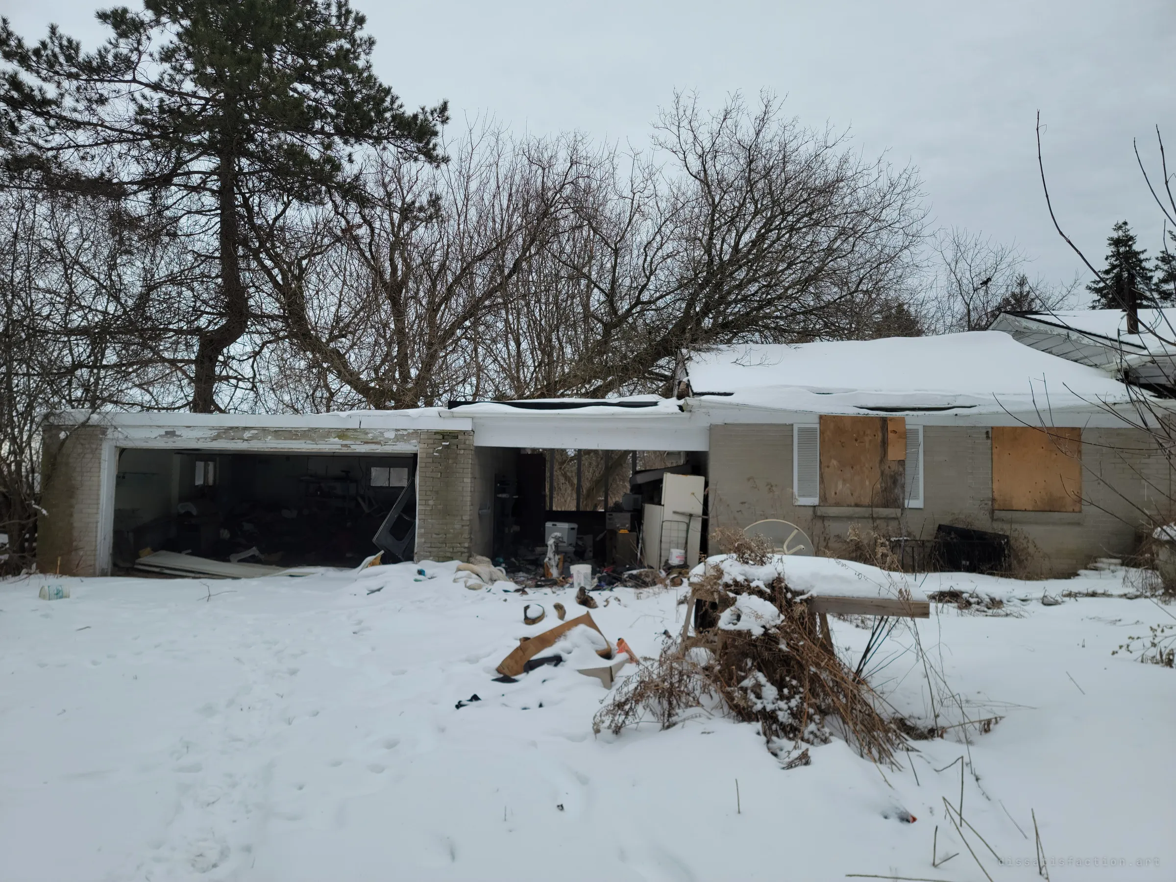 The garage end of the house stands open to the snow, its door gone and junk spilling out across the yard. Boarded windows and a sagging roof show how long it has been abandoned.