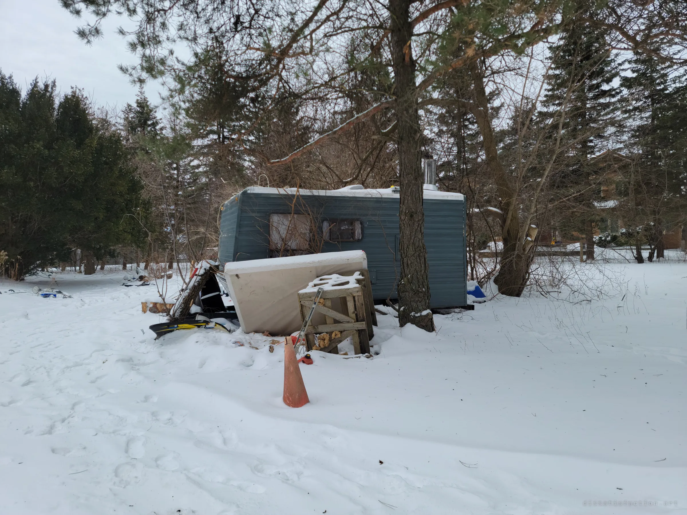 An old camper trailer rots among pines in the snowy yard, a mattress and broken crate propped against it beside an orange traffic cone. It hints that someone lived out here after the house was shut.