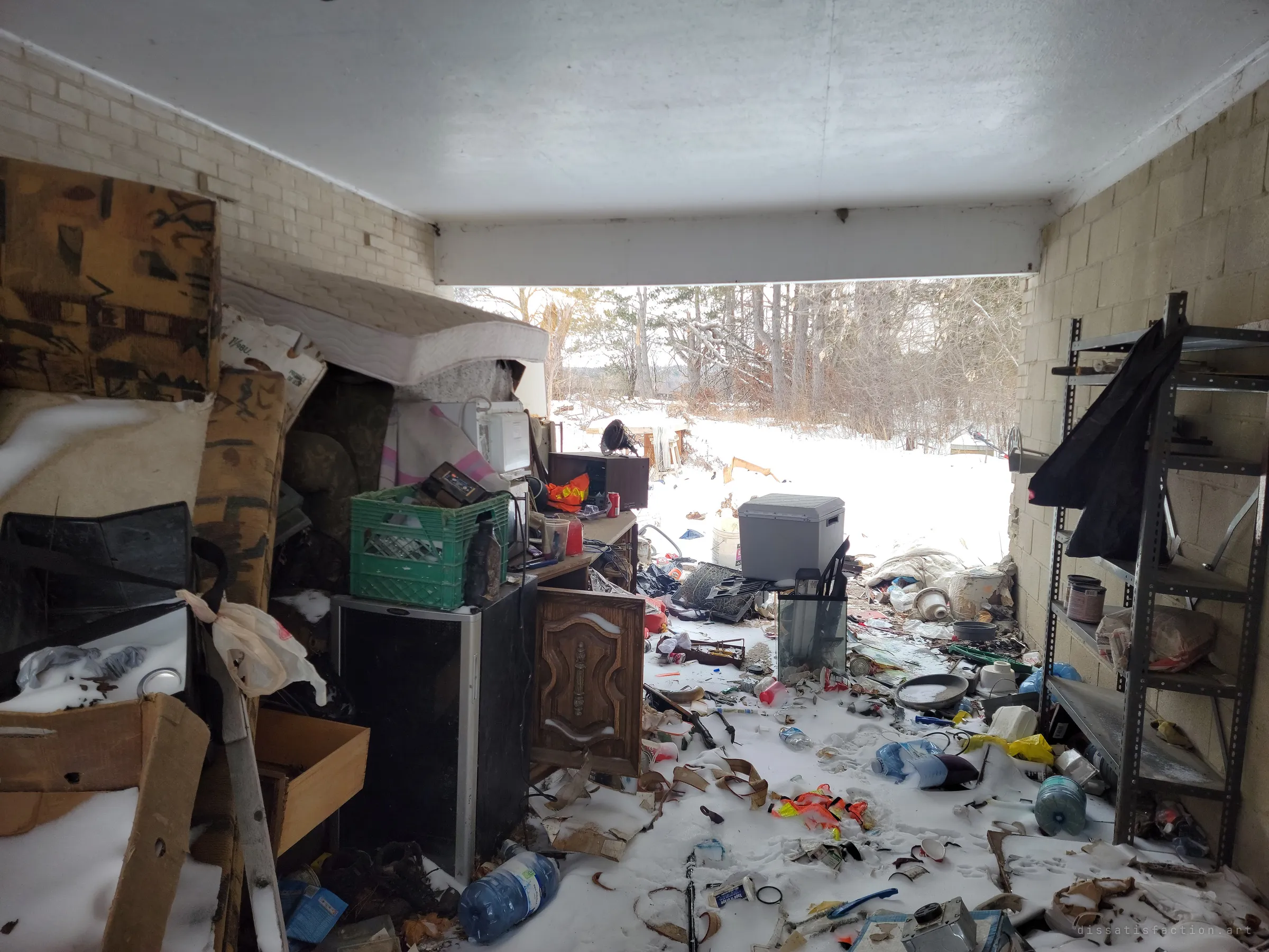 Looking out through the carport, piled furniture, mattresses and shelving give way to snow drifted over scattered trash. The open side frames the woods behind the house.