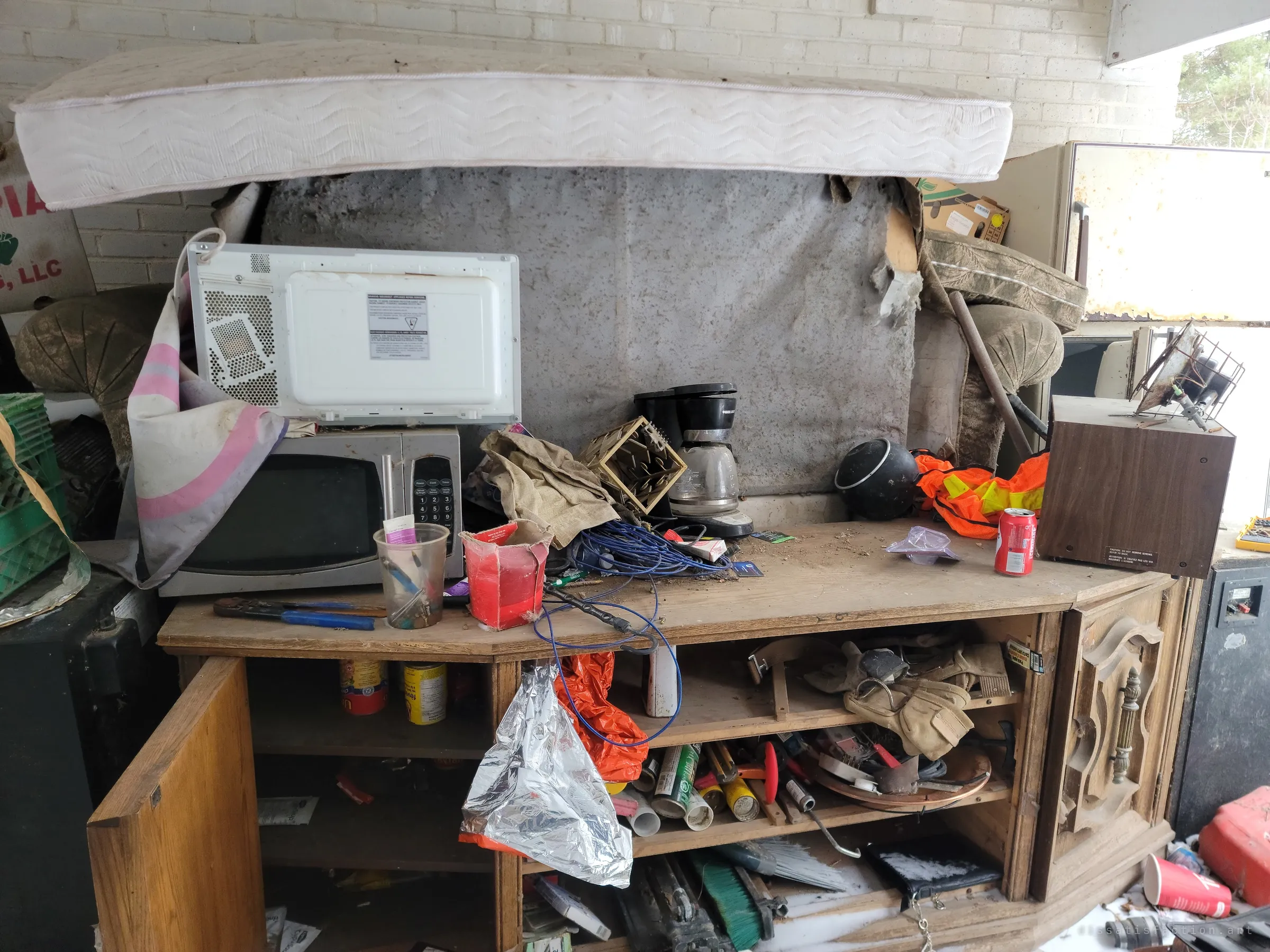 A wooden TV cabinet has become a workbench piled with two microwaves, a coffee maker, tools and a soda can, with a mattress laid across the top. The clutter shows everyday life abandoned mid-use.
