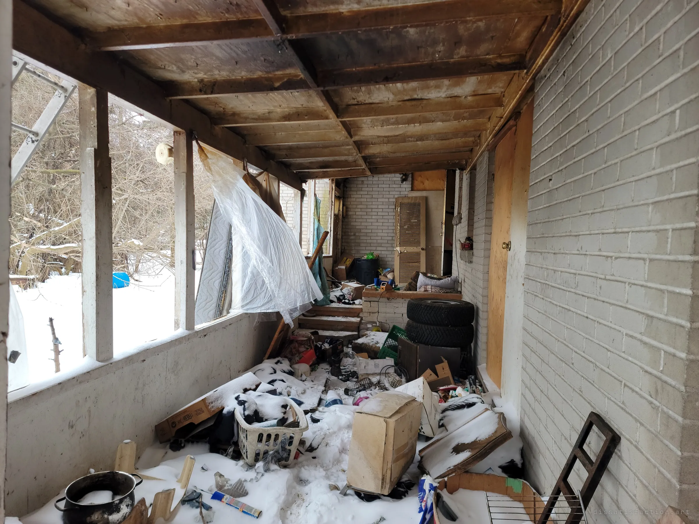 A narrow enclosed porch runs along the brick wall, choked with snow, boxes, a laundry basket, tires and a burnt pot. Torn plastic sheeting hangs from the open window frames.