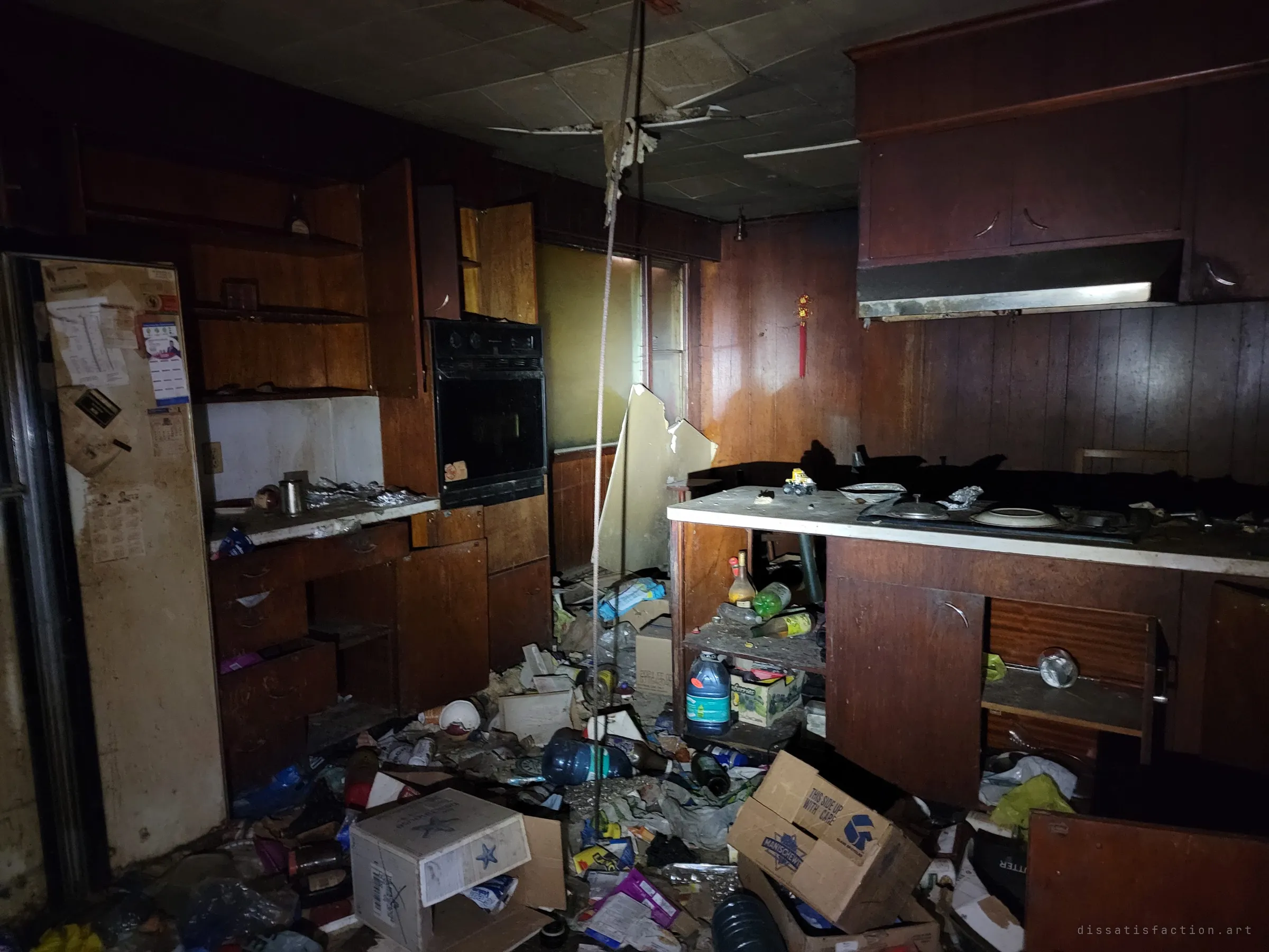 A flashlight picks out a dark wood-panelled kitchen with cabinet doors hanging open and the floor buried in bottles and boxes. Faded papers and calendars still cling to the fridge door.