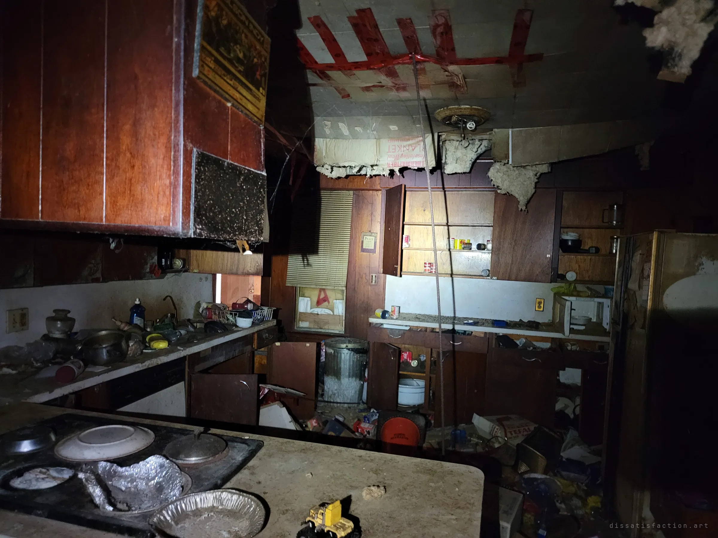 From behind the stovetop, the kitchen ceiling sags open with insulation spilling through, crossed with strips of red tape. Dirty plates and a toy truck sit on the counter where they were left.