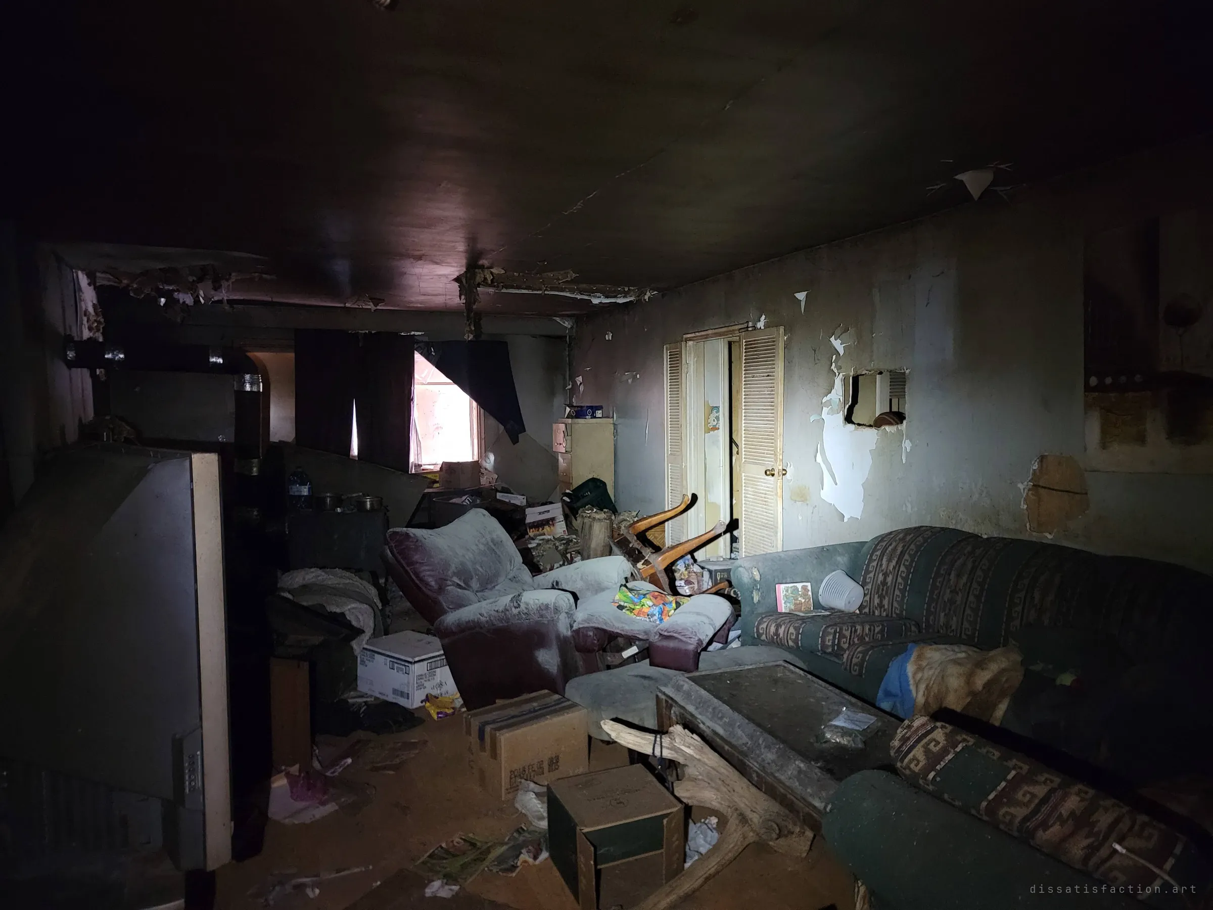 A dim living room is packed with a recliner, patterned couches, boxes and an old rear-projection TV, paint peeling off the walls. A single curtained window lets in a sliver of daylight.