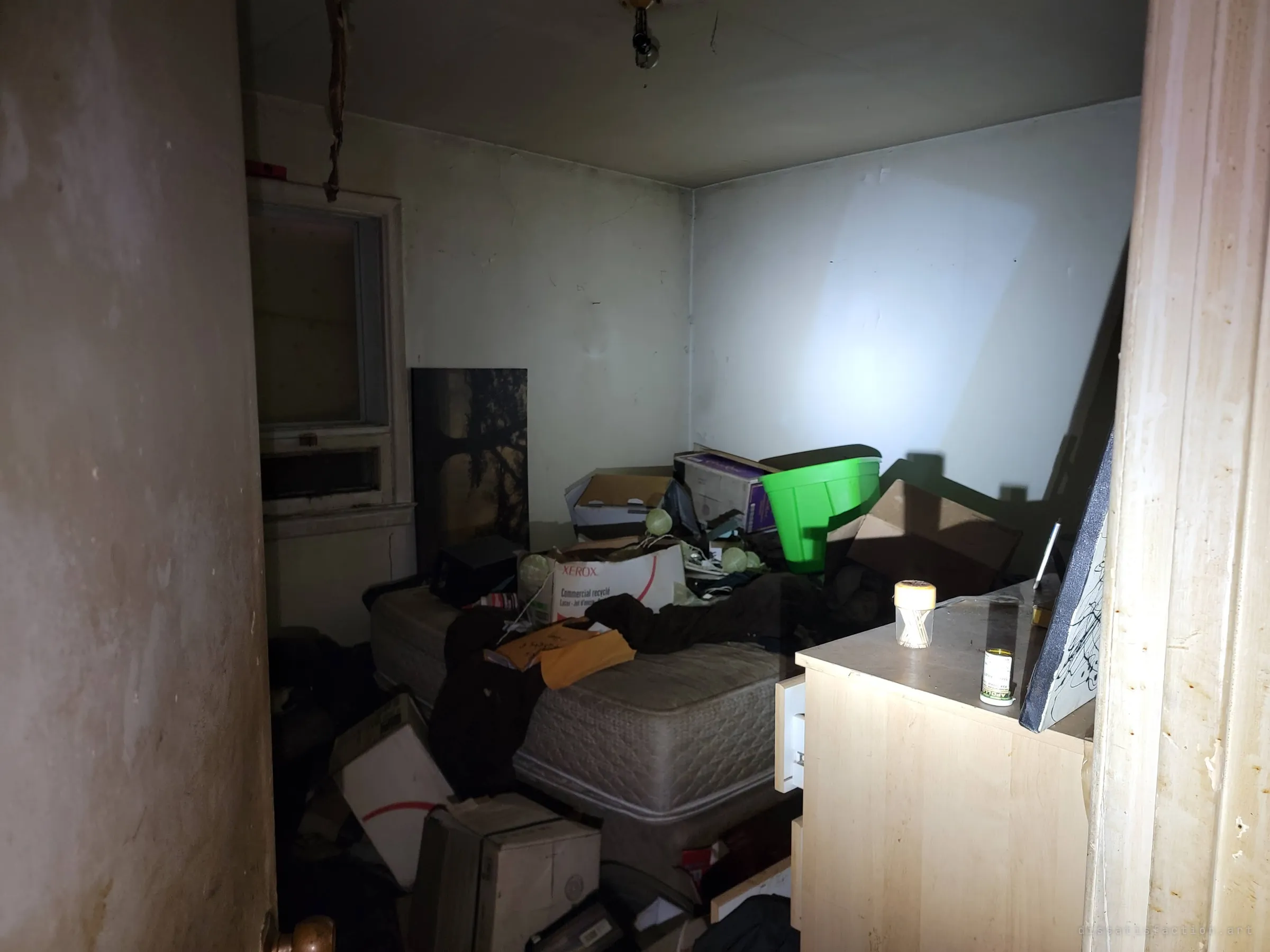 A small bedroom is heaped with boxes, clothes and envelopes on a bare mattress, a bright green bin perched on top. The flashlight leaves the boarded window in shadow.