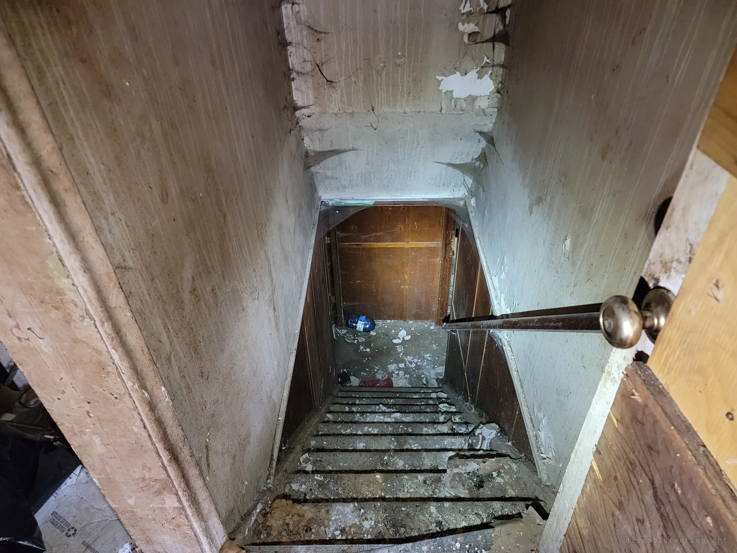 Looking down a narrow staircase to the basement, the steps are littered with fallen plaster and the walls are streaked and peeling. A wood-panelled door waits at the bottom.