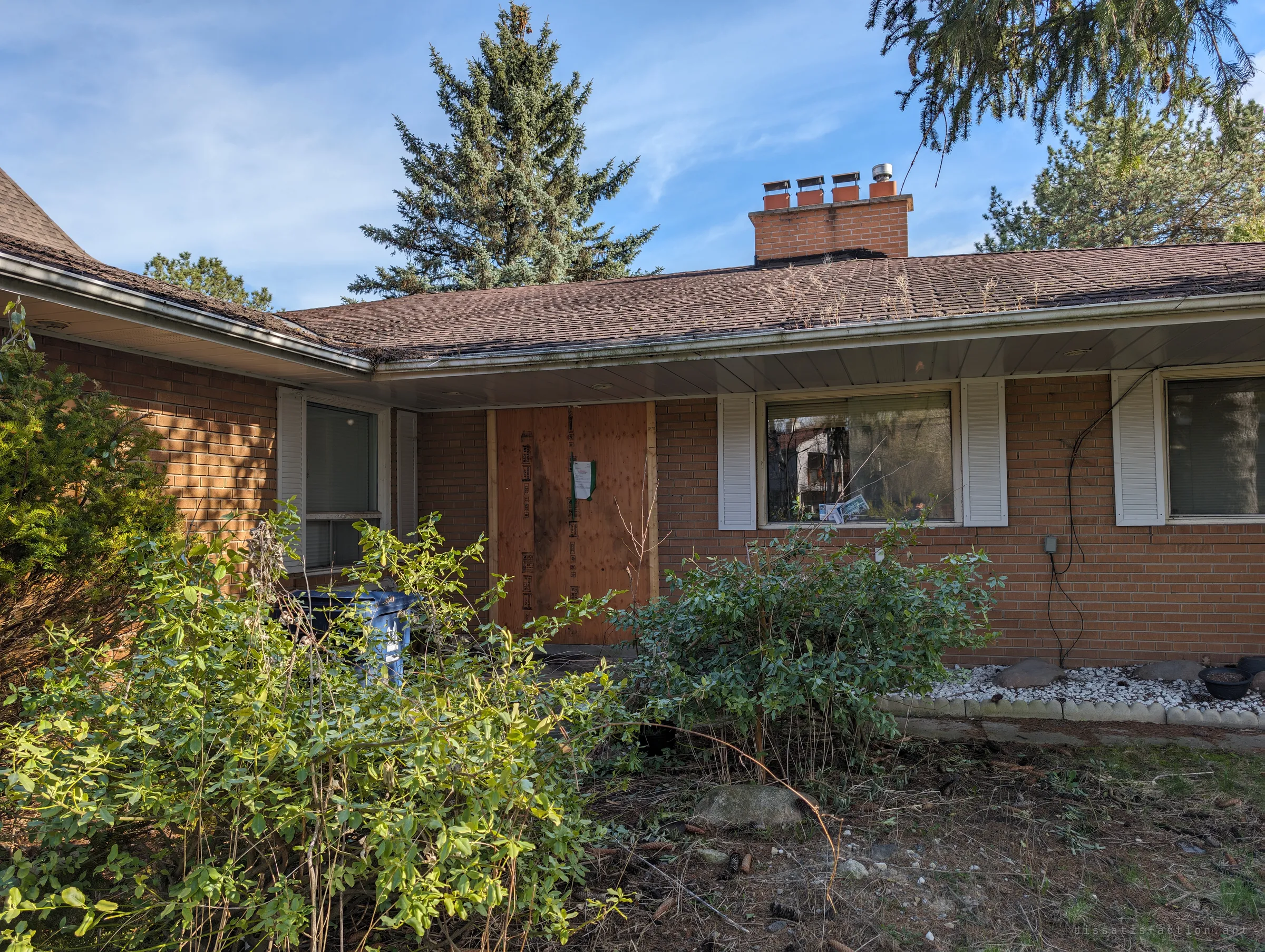 The front of an abandoned brick bungalow, its door boarded with plywood and an official notice, half hidden behind overgrown shrubs. Sets the scene: a family home sealed up and left waiting.