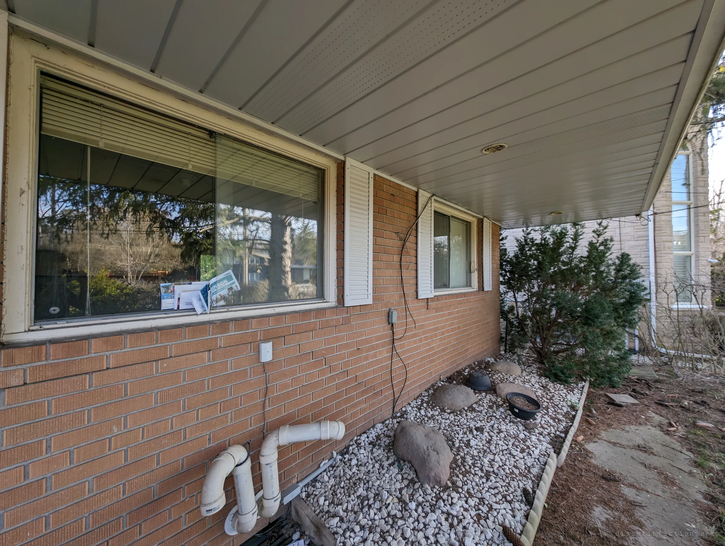 A wide view along the house's brick side under the eaves, unopened mail piled against the window above a bed of white stones. The uncollected post shows how long nobody has been home.