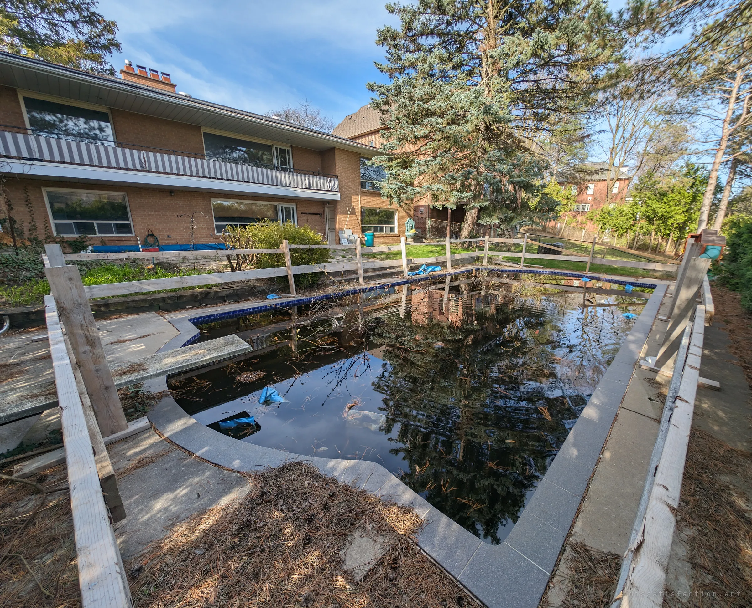 The backyard pool, fenced off with rough lumber and filled with dark still water, pine needles and debris, the two-storey house behind it. The flooded pool is the clearest sign the property has been left to rot.