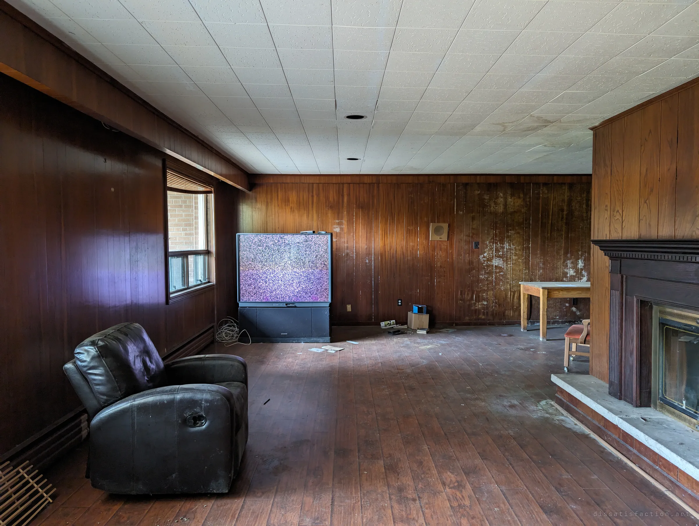 A long wood-panelled basement room with a lone leather recliner facing a big rear-projection TV showing static, a fireplace at right. The glowing static makes the empty room feel like someone just left.