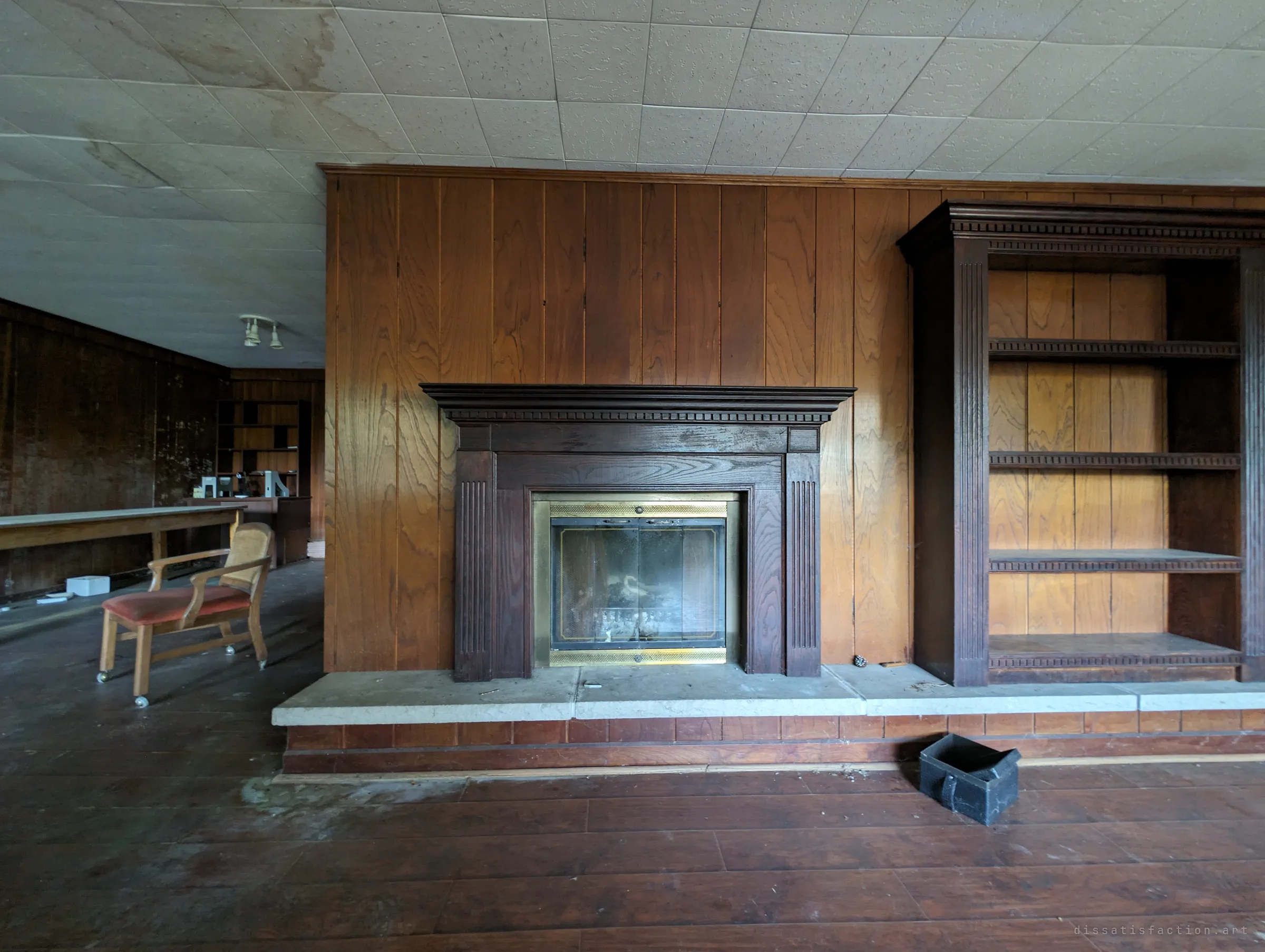 A dark wood fireplace with a brass screen and built-in shelving set into a panelled wall, a rolling chair and long table beyond. The cold hearth anchors the abandoned family room.