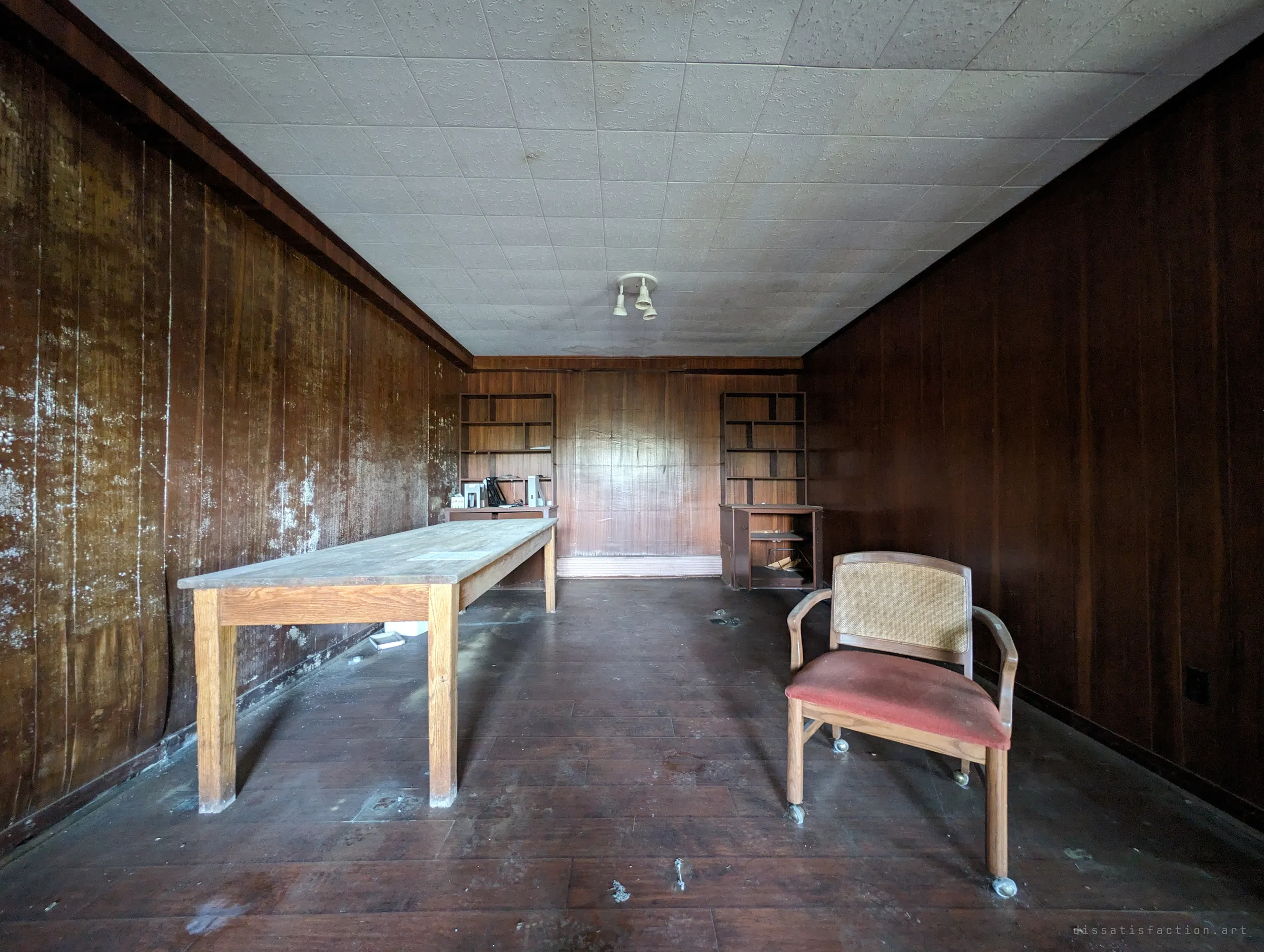 A narrow panelled room with a long bare wooden table and a single red-seated chair, empty shelves at the far end and mould spotting the walls. The symmetry and emptiness give it a waiting-room stillness.