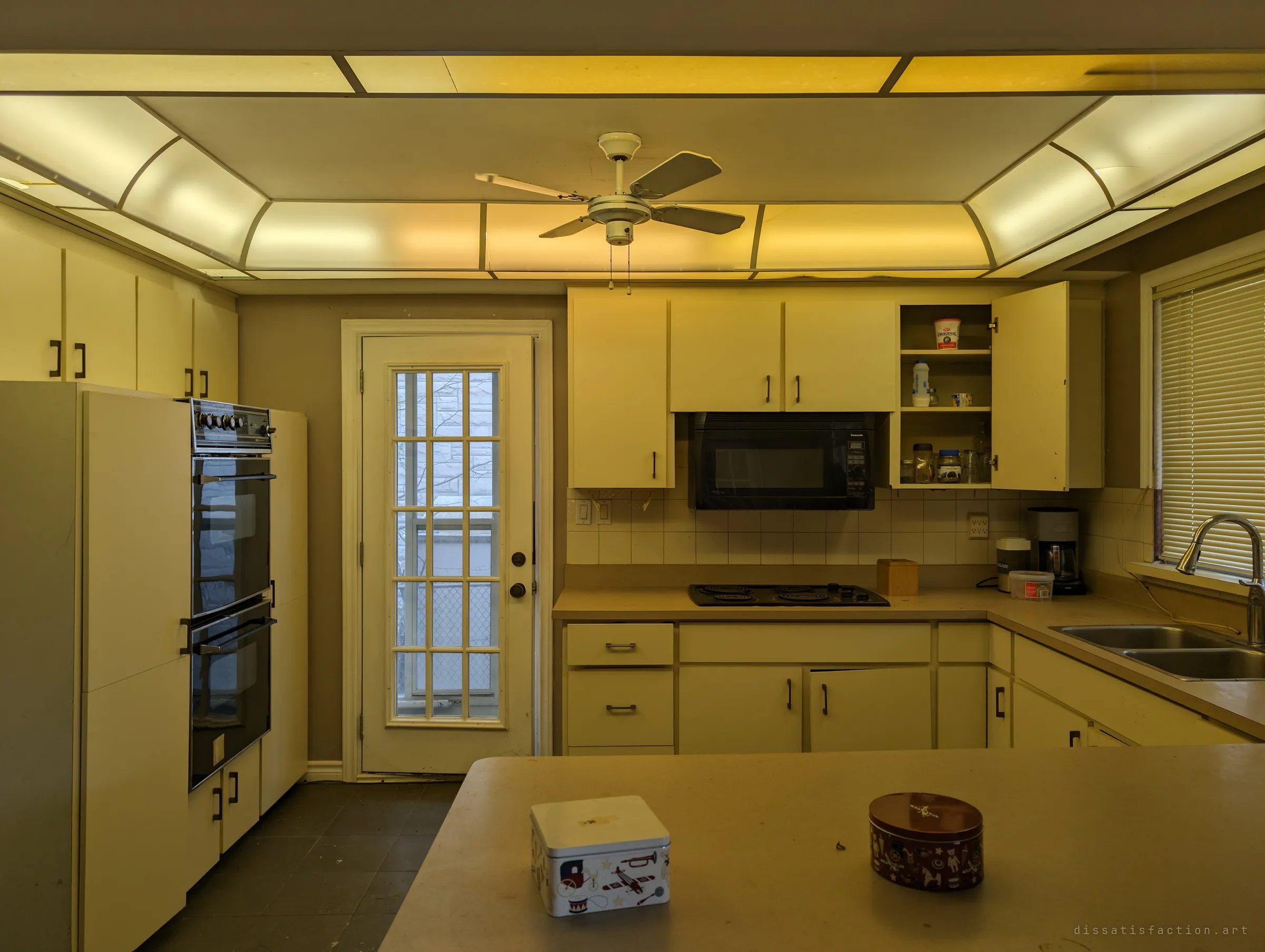 A dated cream kitchen under a glowing yellow ceiling light panel, a ceiling fan above and cabinets still stocked with jars. Two decorative tins left on the counter make it feel only just abandoned.
