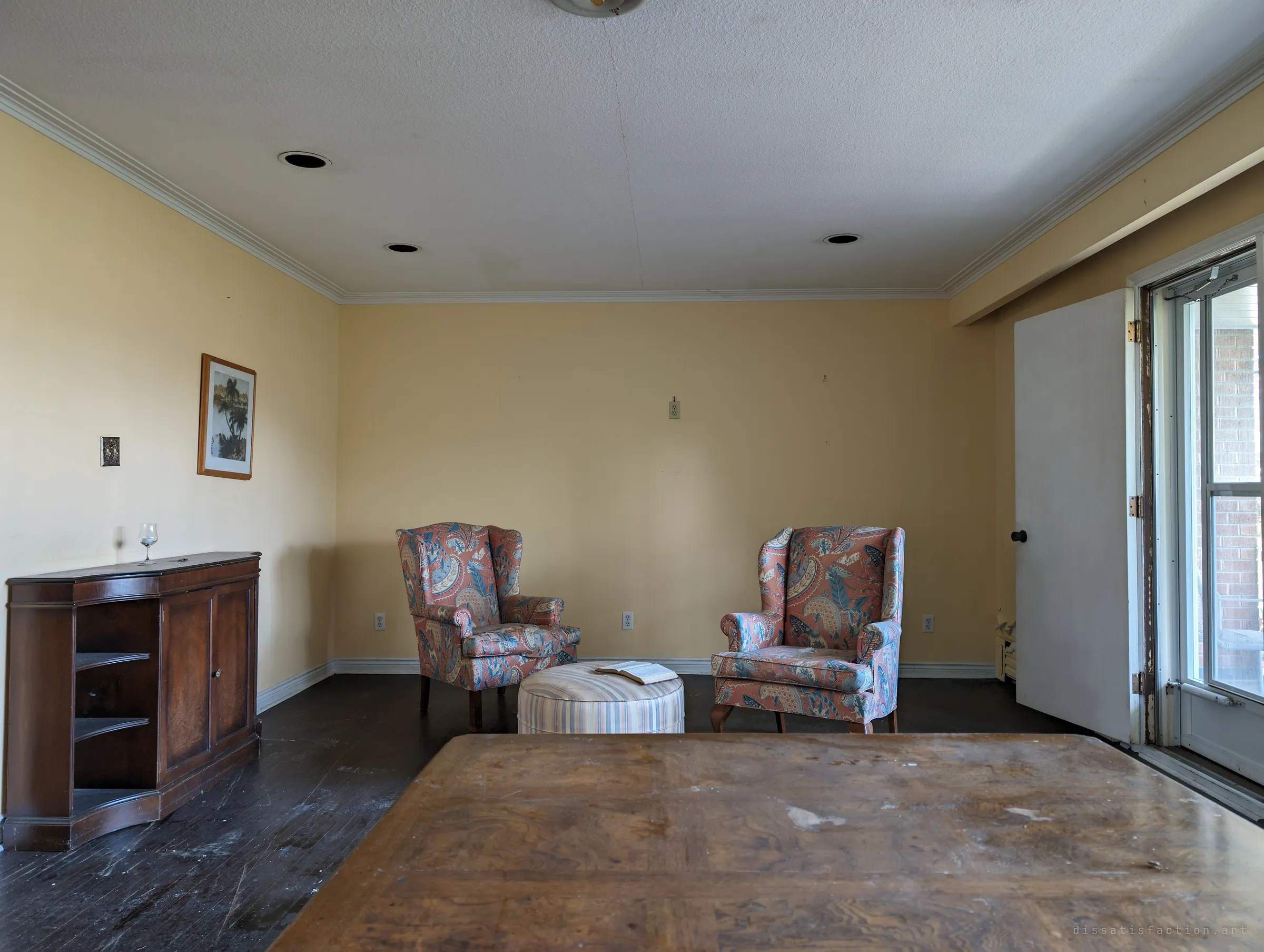 Two floral wingback chairs face each other across a striped ottoman with an open book, a sideboard with a lone wine glass at left and a worn table in the foreground. The staged pairing reads like a conversation that never finished.