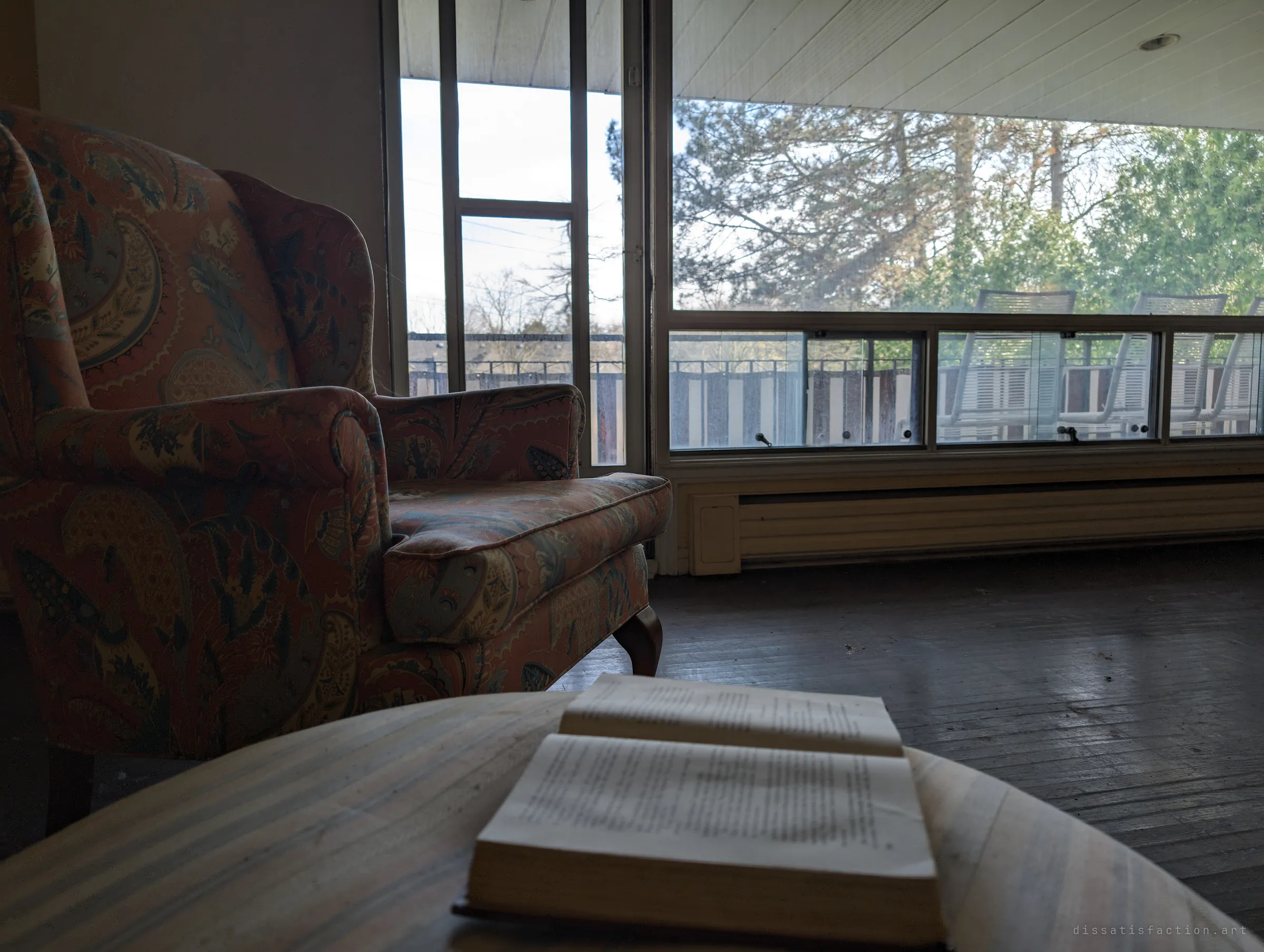 A paisley wingback armchair beside an open book on a side table, facing a wall of windows onto an empty balcony. The room looks paused mid-afternoon, as if the reader just stepped out.