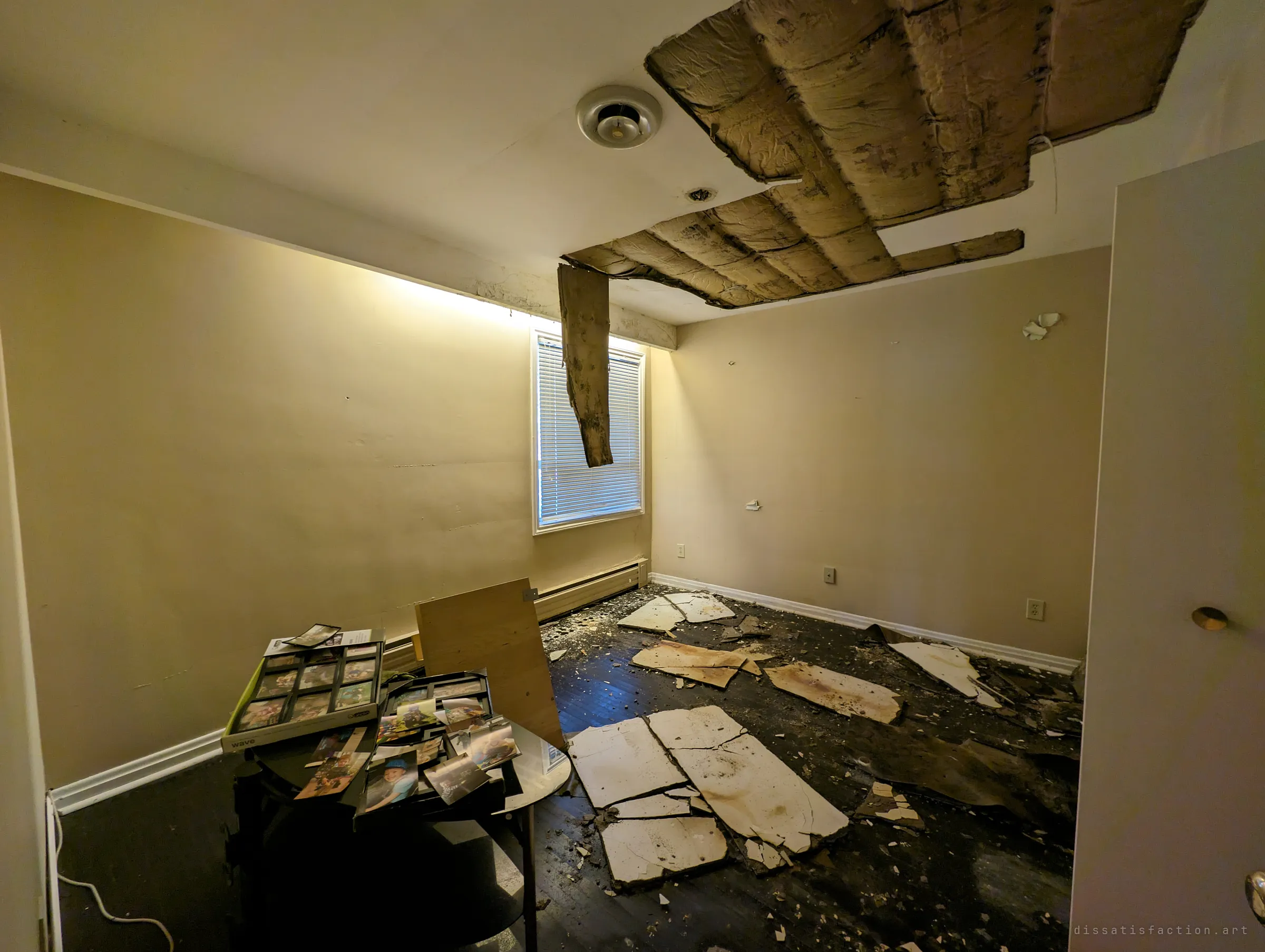 A small bedroom where the ceiling has given way, insulation sagging and drywall slabs scattered across the dark floor. Family photo frames still sit piled on a table in the corner.