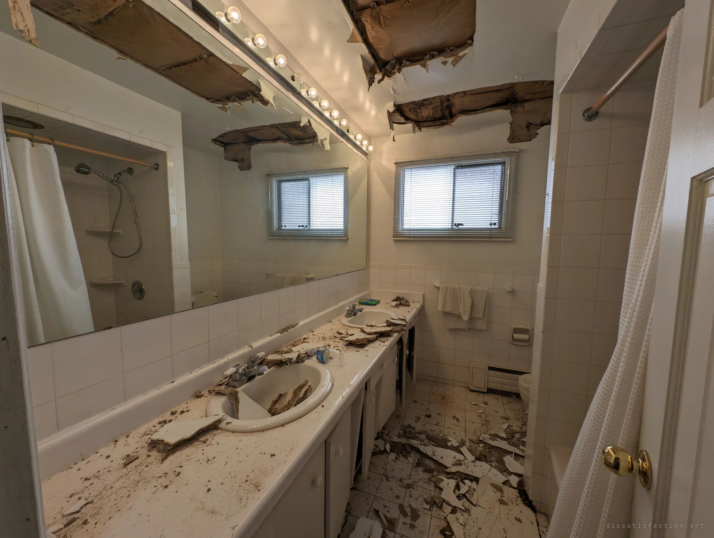 A long double-sink bathroom with a vanity mirror and bulb strip, the ceiling torn open above it. Plaster and debris cover the counter, the sinks and the tiled floor.