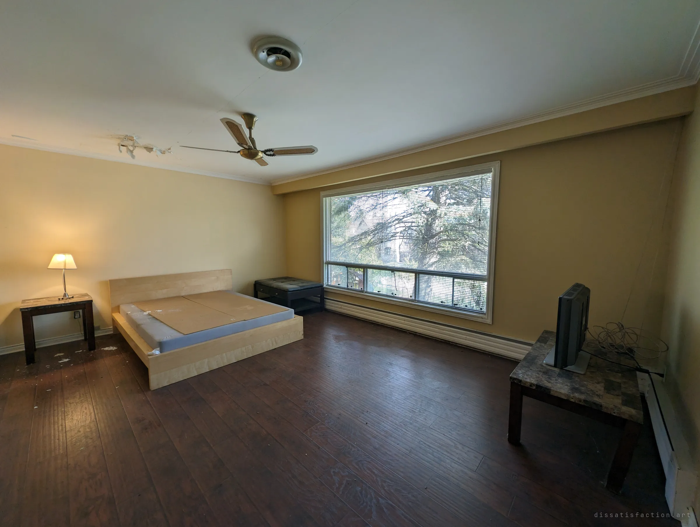 A spacious bedroom with a bare bed frame, a lamp still lit on the nightstand, a ceiling fan and a small TV on a side table. Apart from a patch of peeling ceiling it looks almost lived-in.