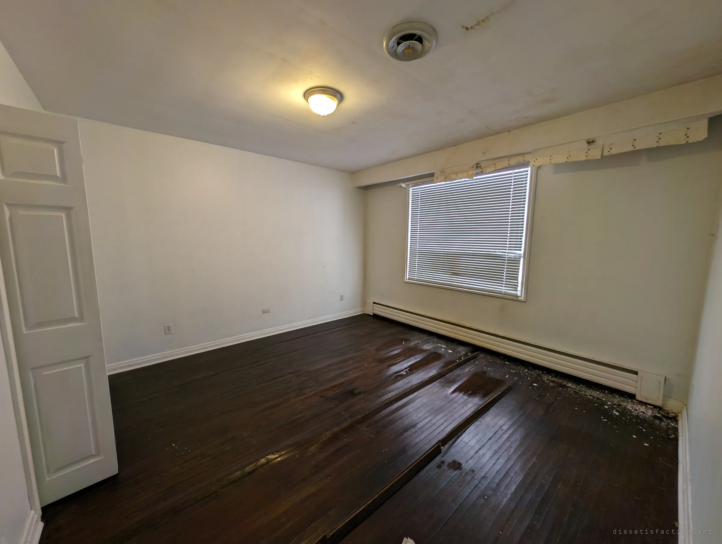 An empty bedroom with dark hardwood floors buckled and lifting from water damage. Stained ceiling and a strip of peeling wallpaper border above the window show the leaks spreading.