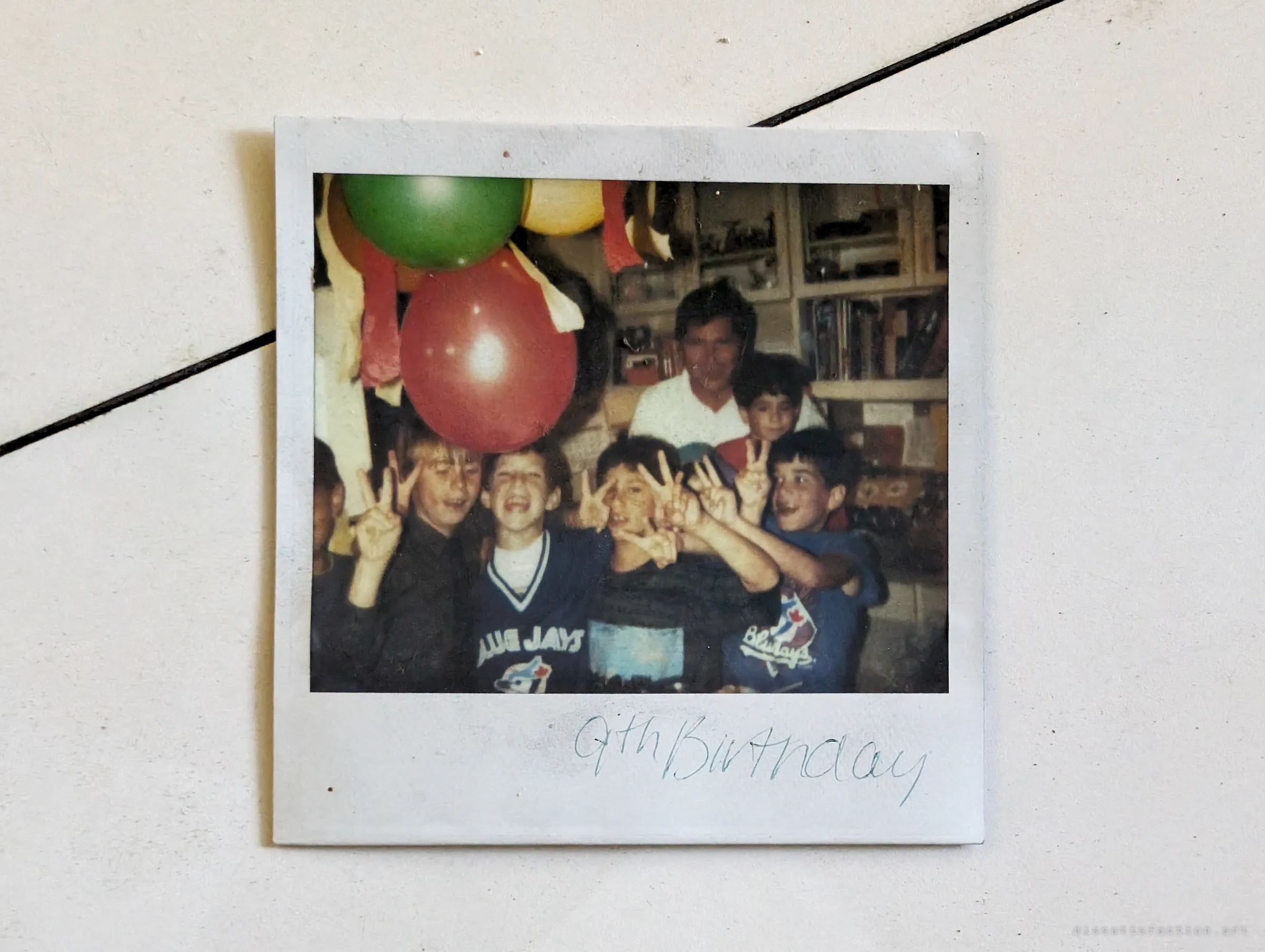An old instant photo left on a tiled surface: a group of kids flashing peace signs at a birthday party under balloons, captioned by hand. A trace of the family who once lived here.