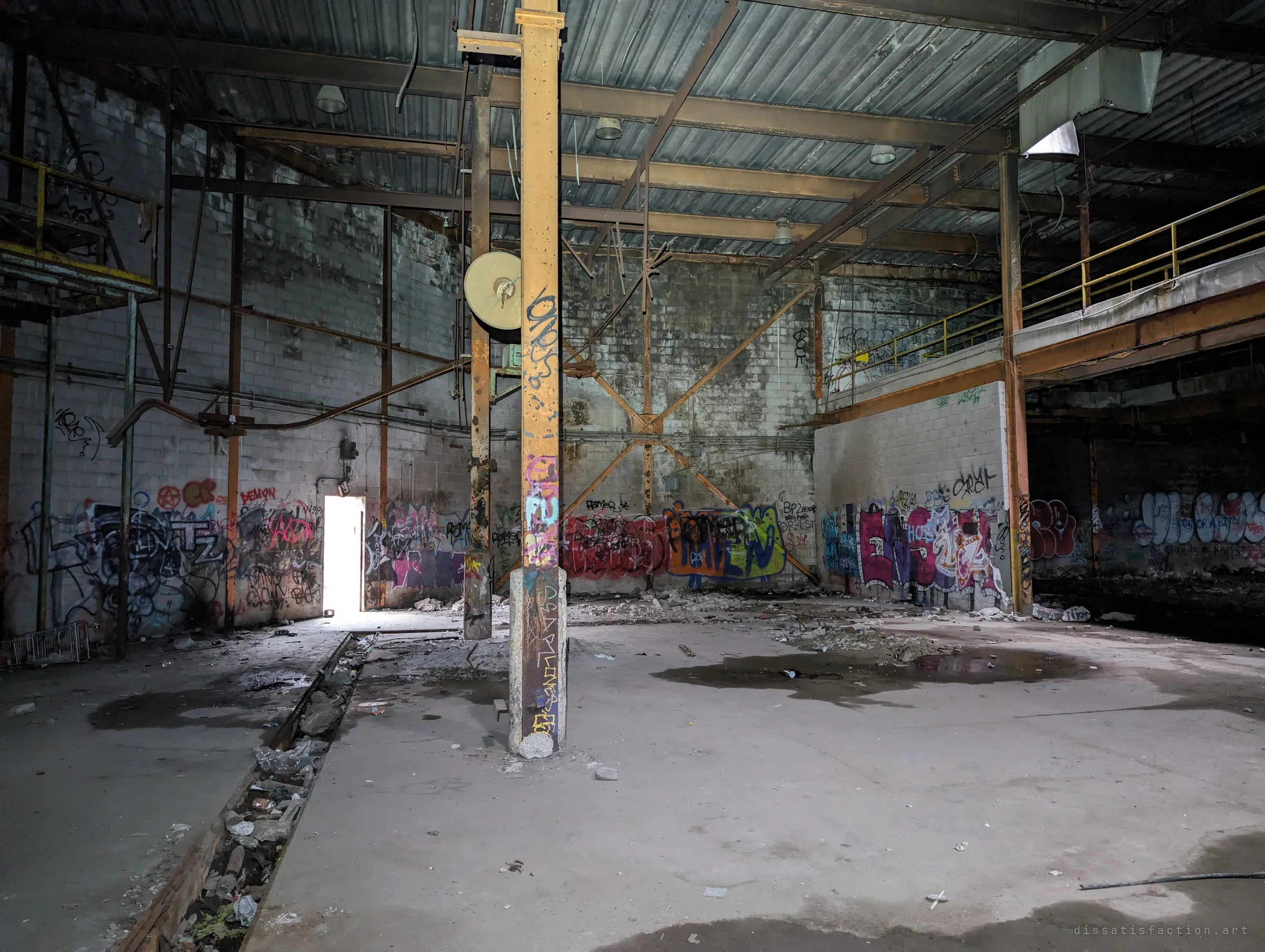A cavernous factory hall with a rusted yellow column in the foreground, a mezzanine walkway on the right and graffiti-covered block walls, lit by a single open doorway. It sets the scale of the abandoned plant.