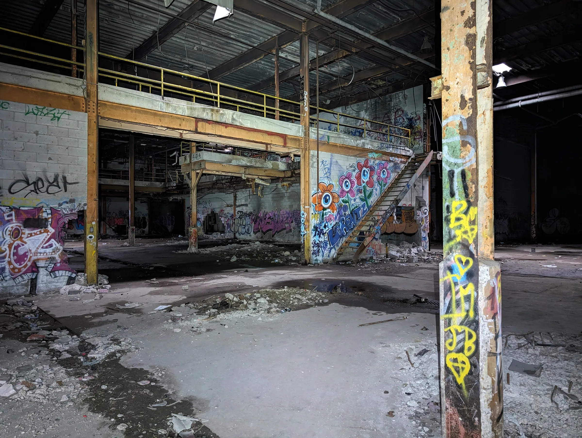 A paint-peeled steel column frames the main floor, where a steel stair climbs to a railed mezzanine past a mural of eyeball flowers. Rubble and puddles cover the concrete.