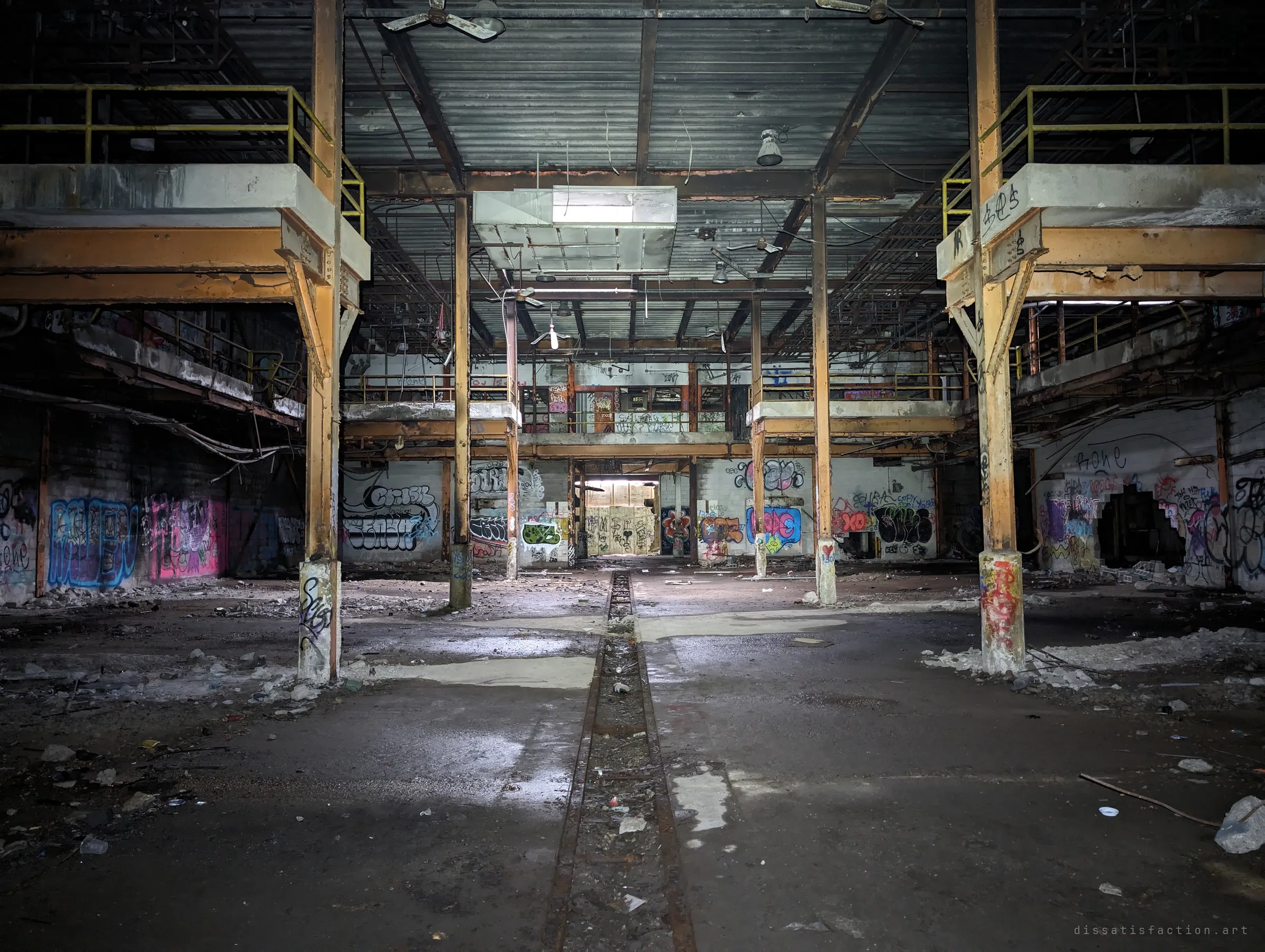 A symmetrical view down the plant's central aisle, with a floor channel running between twin mezzanines toward a boarded, back-lit doorway. Layers of graffiti line every wall.