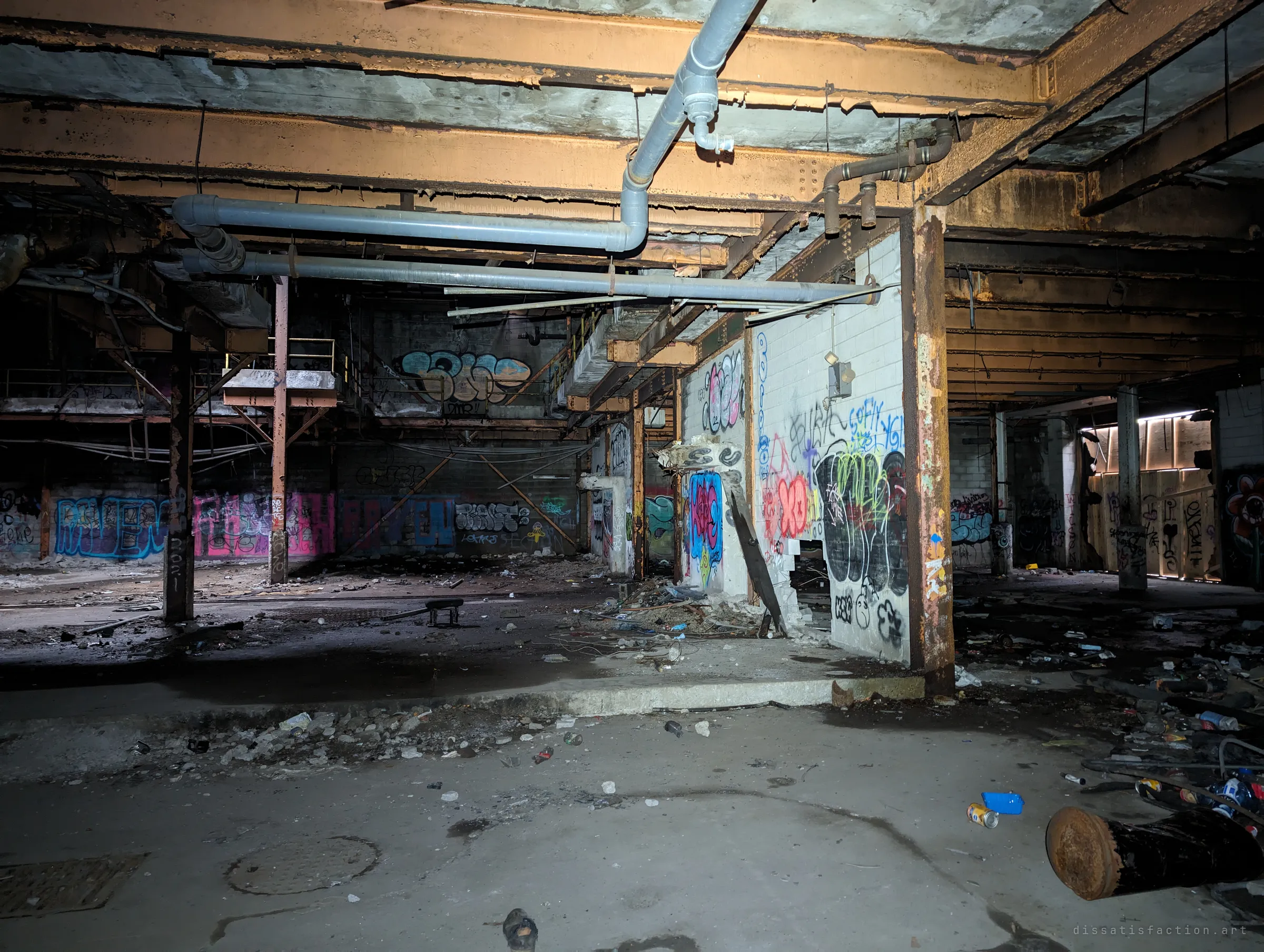 A low-ceilinged lower level under rusted beams and grey pipes, its broken block walls covered in tags and its floor strewn with cans and debris.