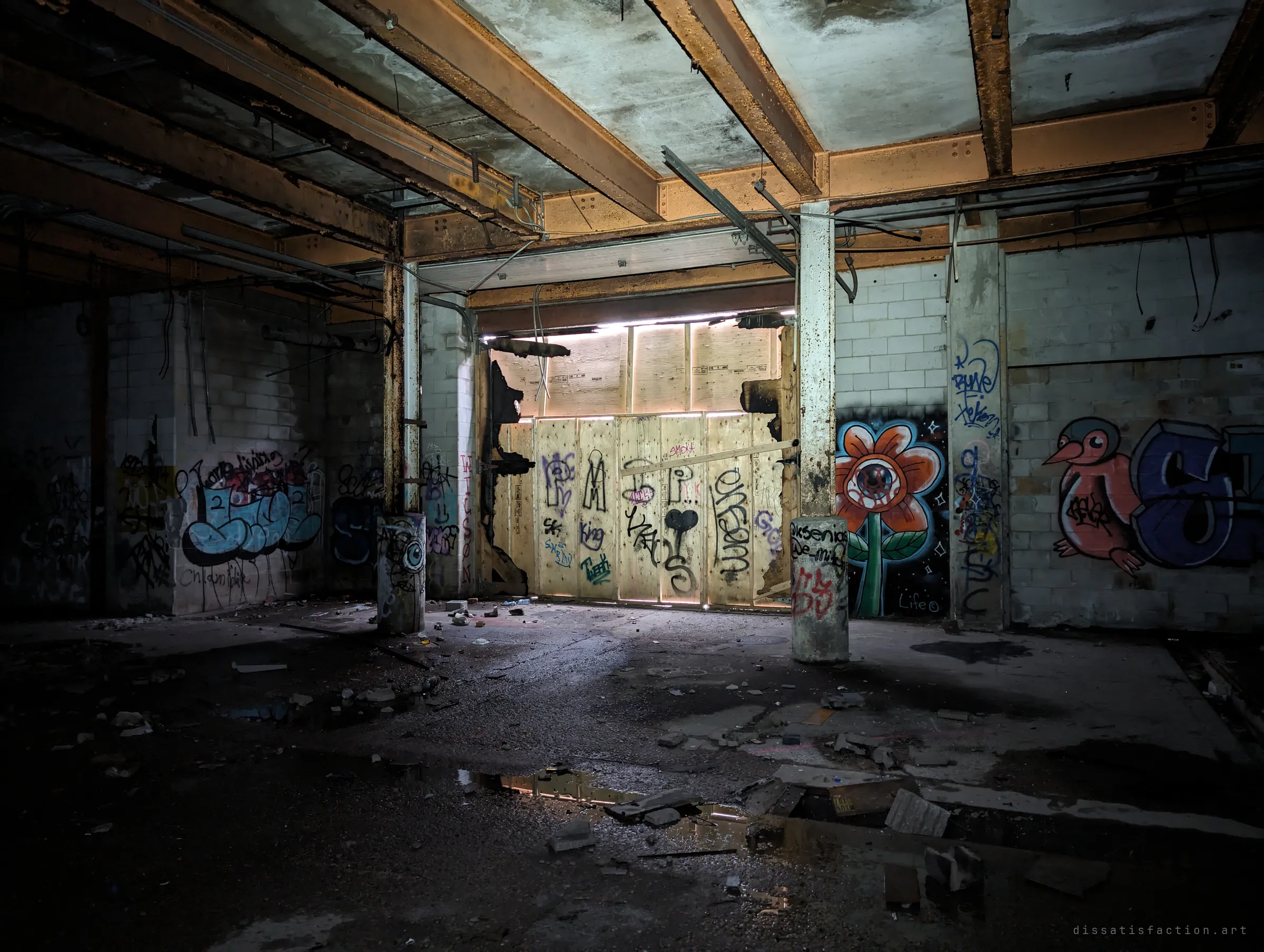 Daylight leaks around a plywood-sealed loading opening, flanked by an eyeball-flower mural and a cartoon bird. Puddles on the floor catch the glow.