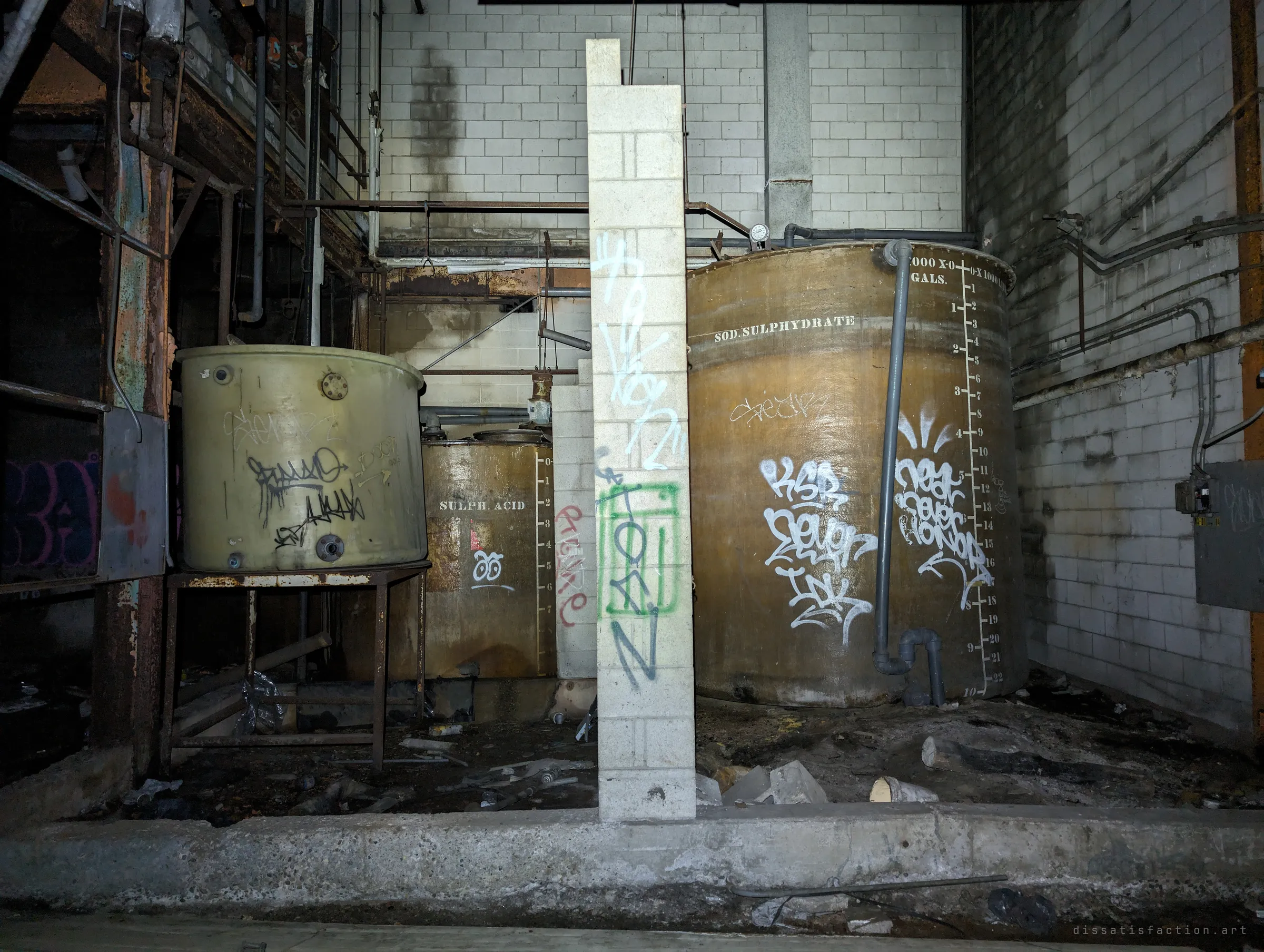 Old process tanks stencilled with chemical names and volume scales stand behind a lone block pillar, all tagged with graffiti. They are what is left of the plant's chemical treatment line.