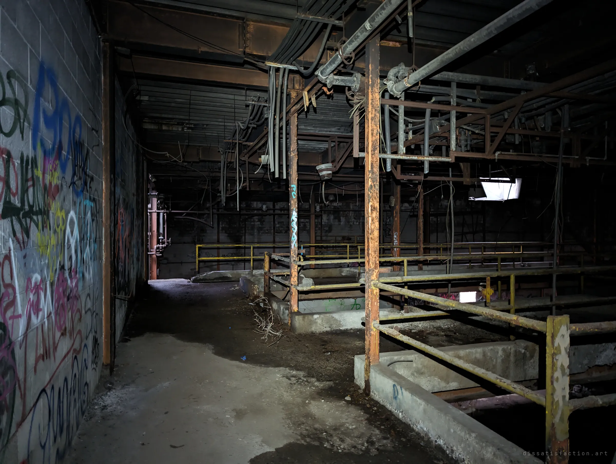 A dark corridor runs beside rows of railed concrete pits under a tangle of hanging cables and pipes. These are likely the old treatment vats of the process floor.