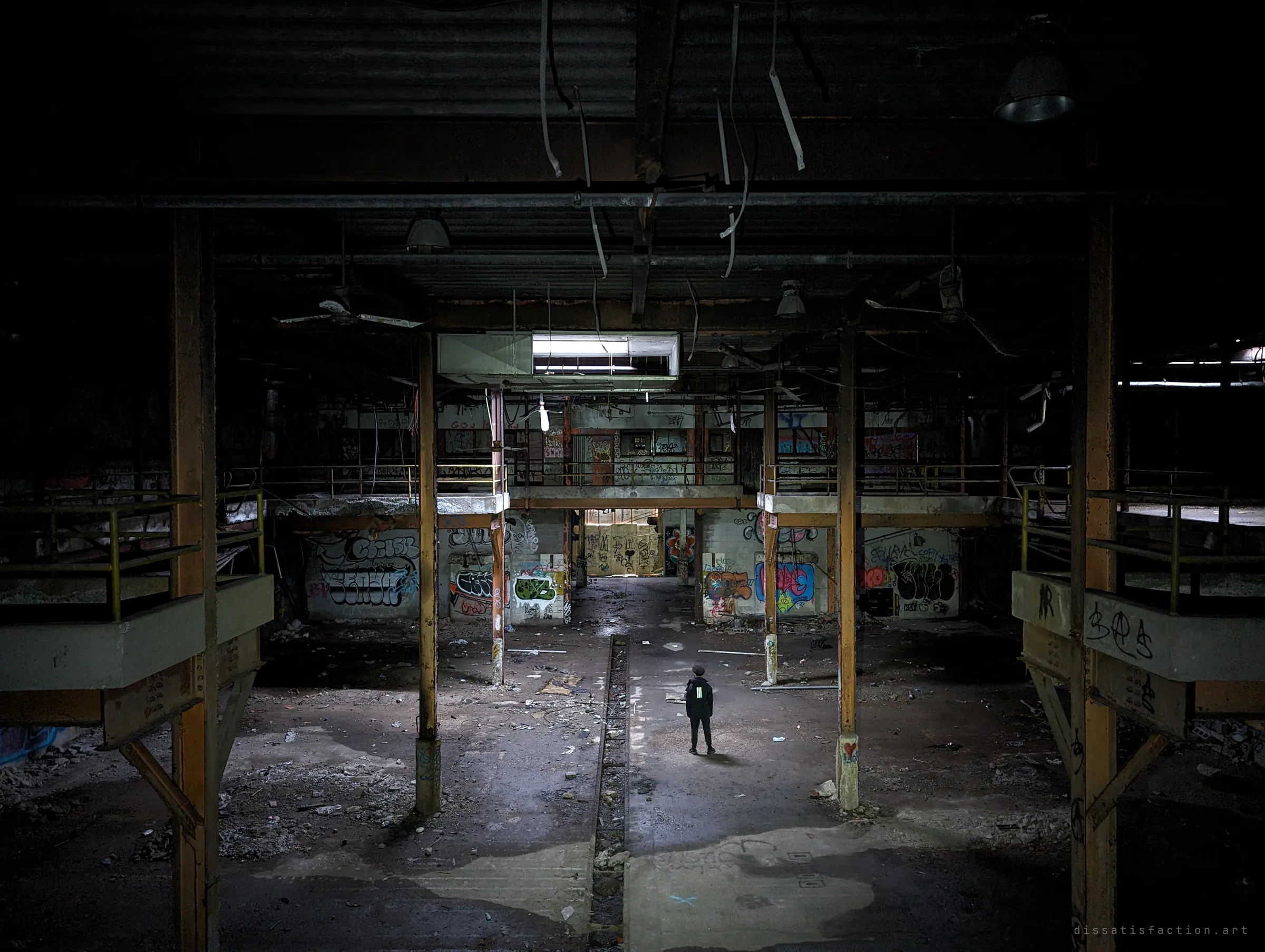 A lone figure stands in the middle of a vast dark factory floor, framed by rusted steel columns, railed mezzanines and graffiti-covered walls under a single skylight. The person gives scale to how big and empty the hall is.