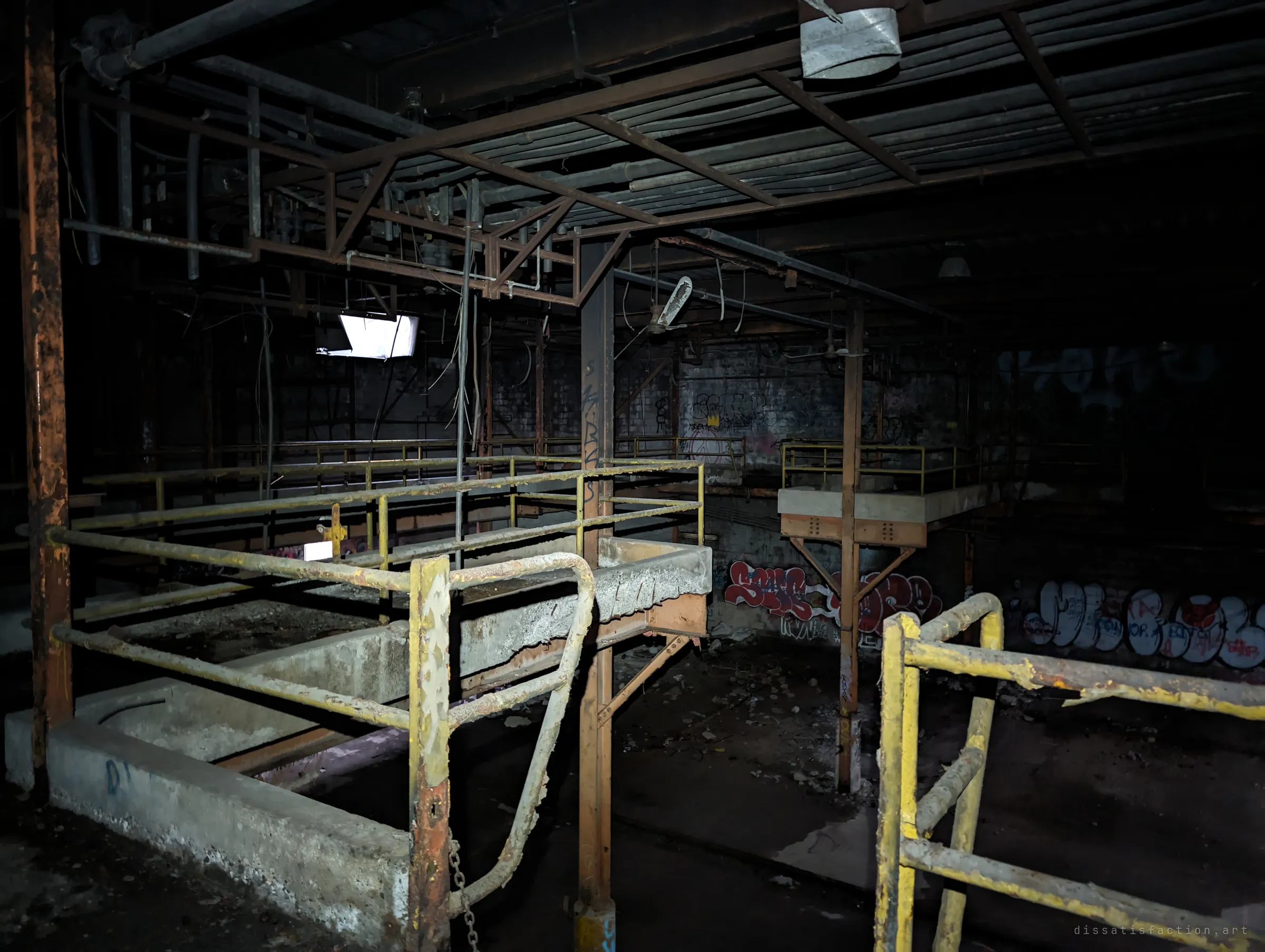 Yellow-painted safety railings wrap around empty concrete process tanks under a tangle of pipes and steel framing, lit by torchlight. The worn rails and old tagging show decades of disuse.
