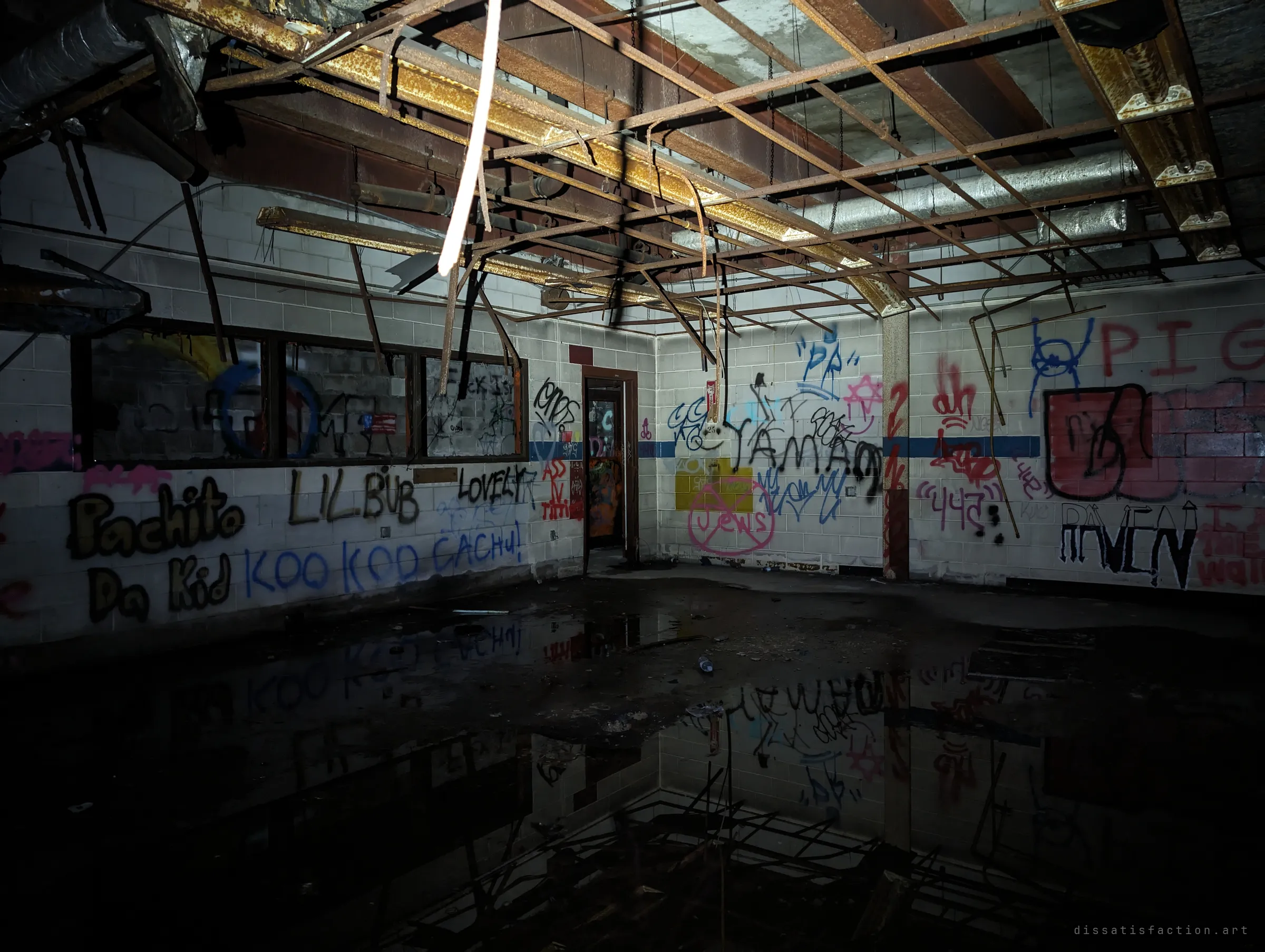 A cinder-block room covered in spray-painted tags, its ceiling grid collapsed and dangling, with a pool of standing water mirroring the walls. The reflection doubles the wreckage of the room.