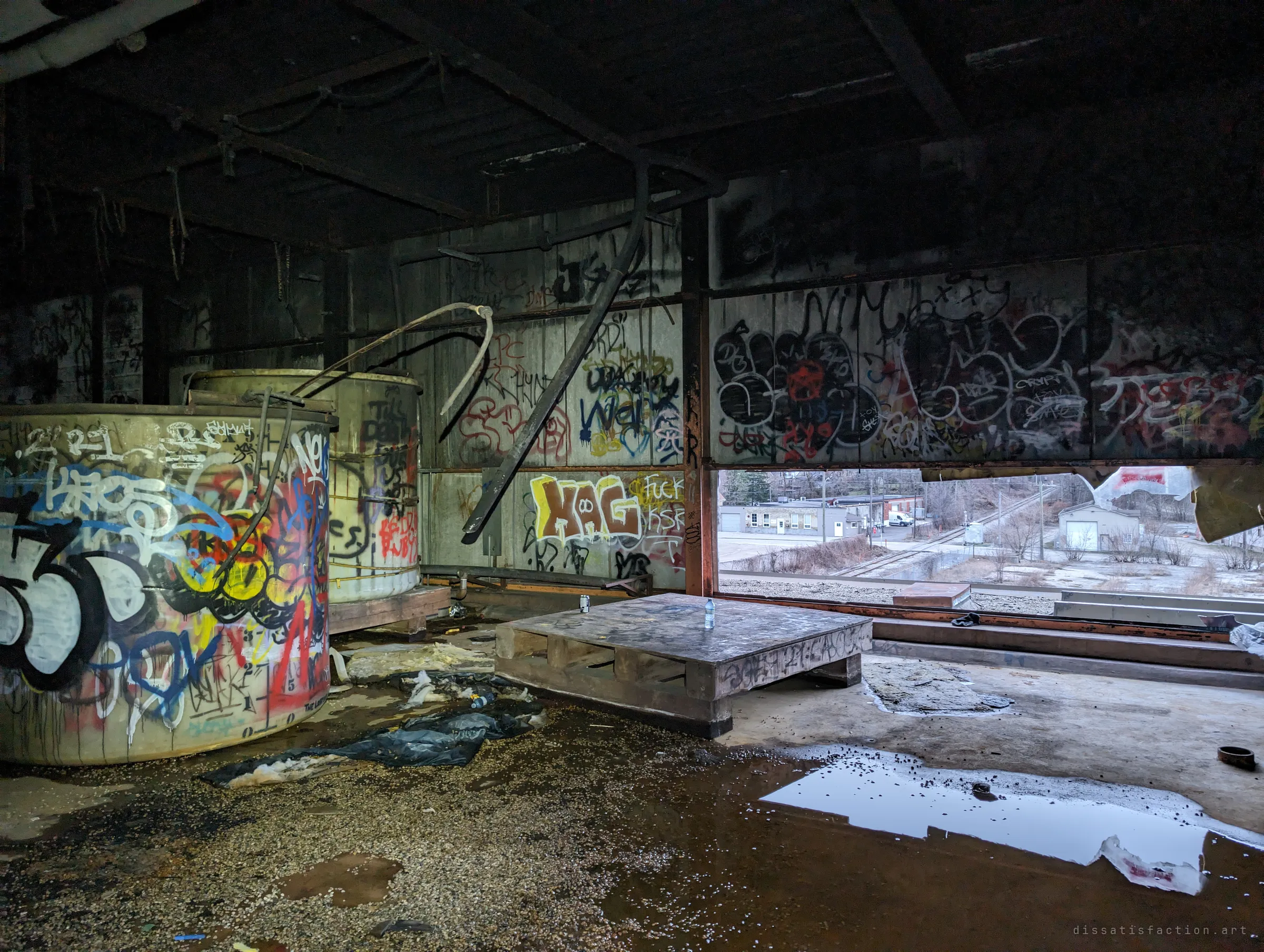 Graffiti-covered storage tanks and a wooden pallet sit in a gutted upper floor whose wall has been torn open to the grey daylight outside. Puddles on the concrete reflect the sky.
