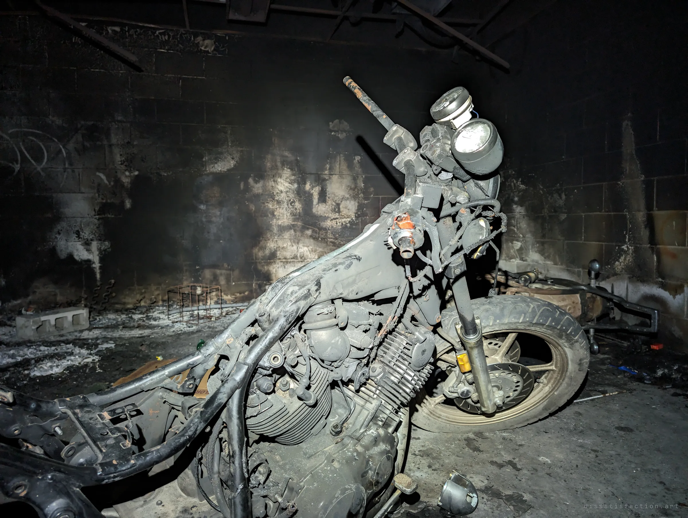 The burnt-out frame of a motorcycle, stripped to engine, forks and gauges, stands against a scorched cinder-block wall. Soot and ash cover everything around it.