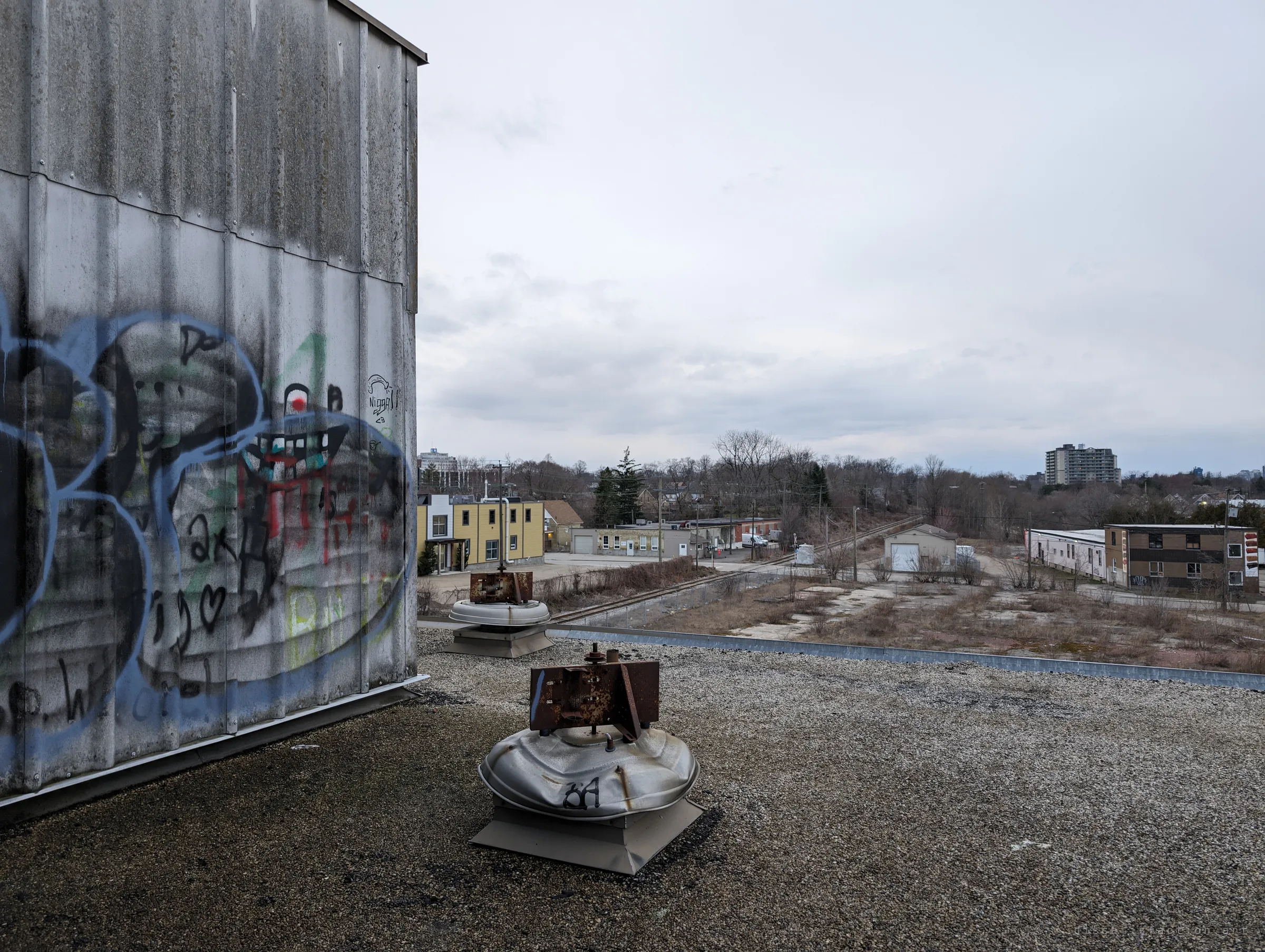 On the gravel roof, a tagged metal wall and rusted rooftop vents look out over an overcast neighbourhood. The open sky is a sharp contrast to the dark interior.