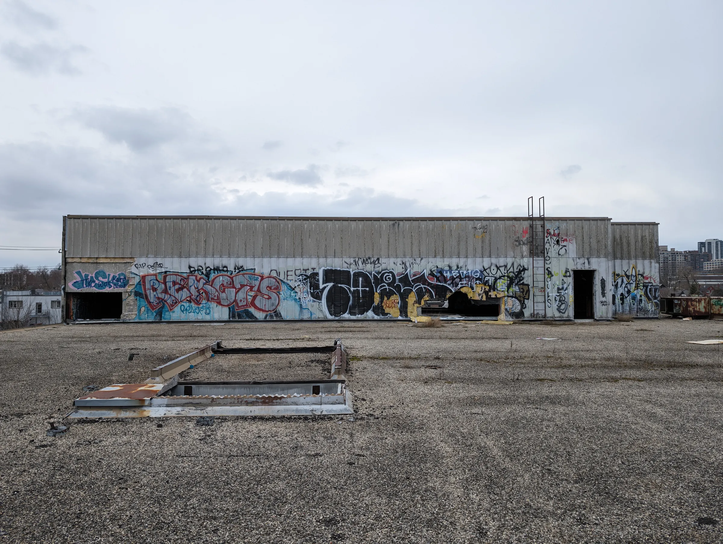 A long, low penthouse structure on the roof is covered end to end in graffiti pieces, with an open skylight frame in the gravel foreground. The flat grey sky makes the colour of the paint stand out.