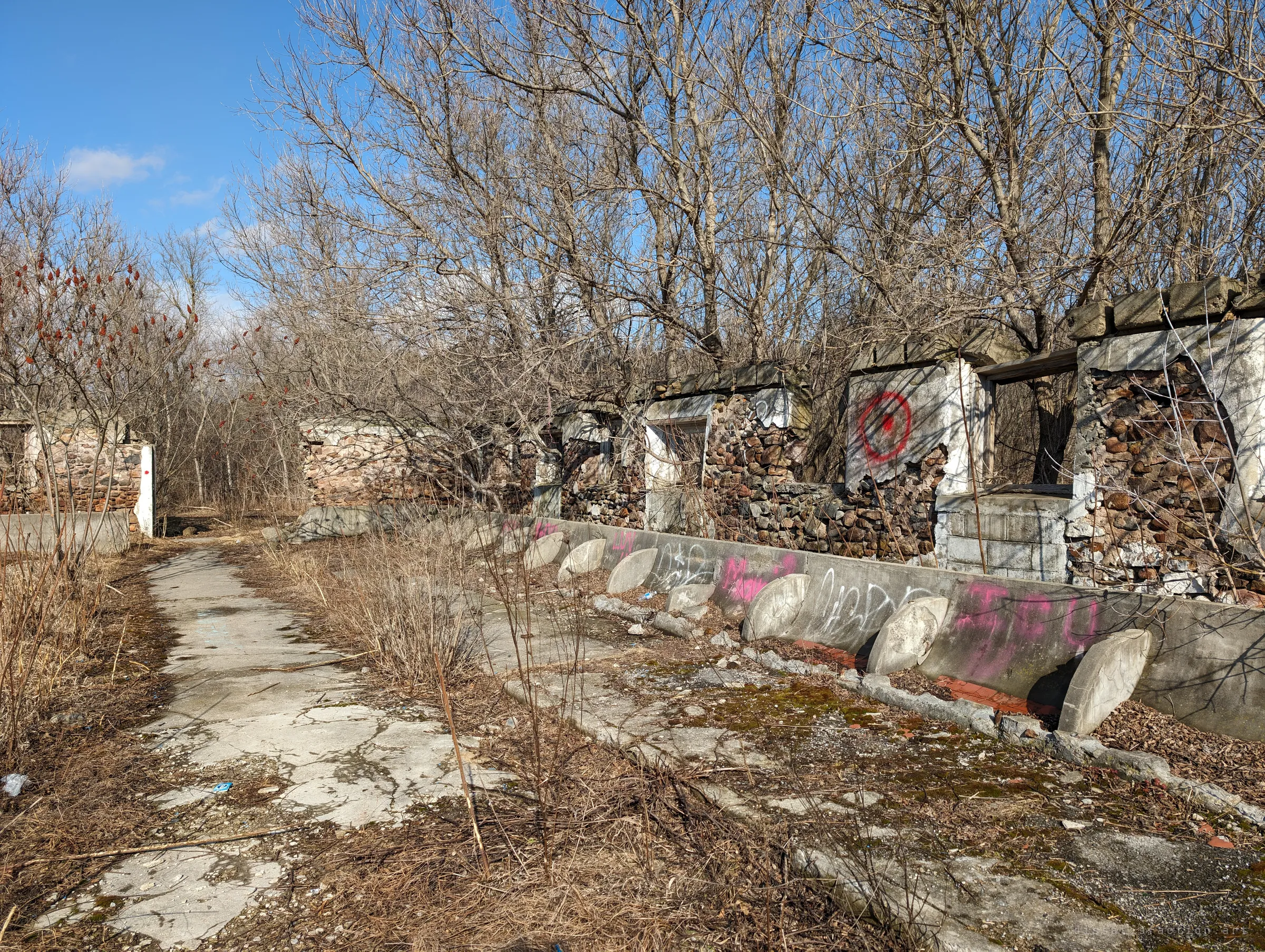 A cracked concrete path runs past a line of fieldstone pens, with fallen round troughs leaning against a graffitied wall and a red target sprayed on one pier. Bare trees have grown up through the ruins.