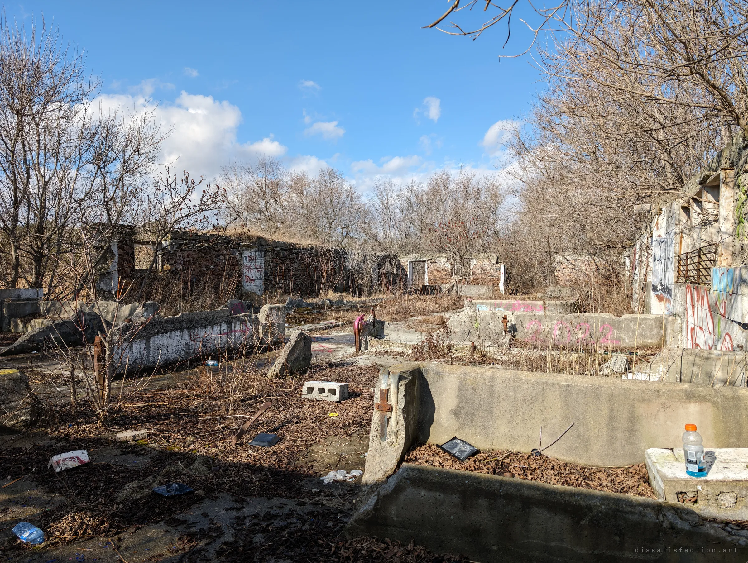 Low concrete walls and broken enclosures littered with cinder blocks, bottles and rubbish, with a stone building and graffiti-covered walls beyond. It shows how the site has become a hangout.